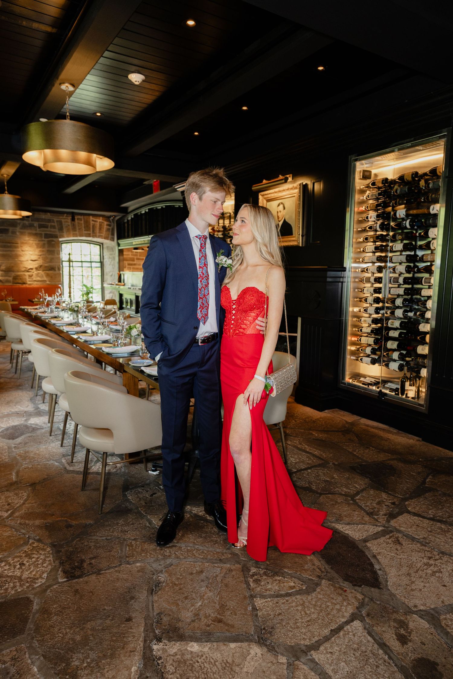 Elegant couple posing in upscale restaurant, man in navy suit, woman in red gown, wine cellar backdrop.
