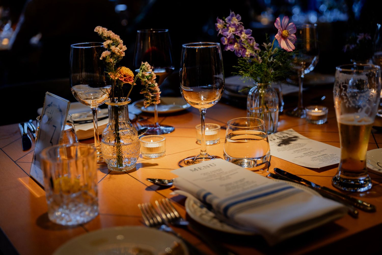 Elegant restaurant table setting with wine glasses, floral arrangements, silverware, and menus in warm candlelight.