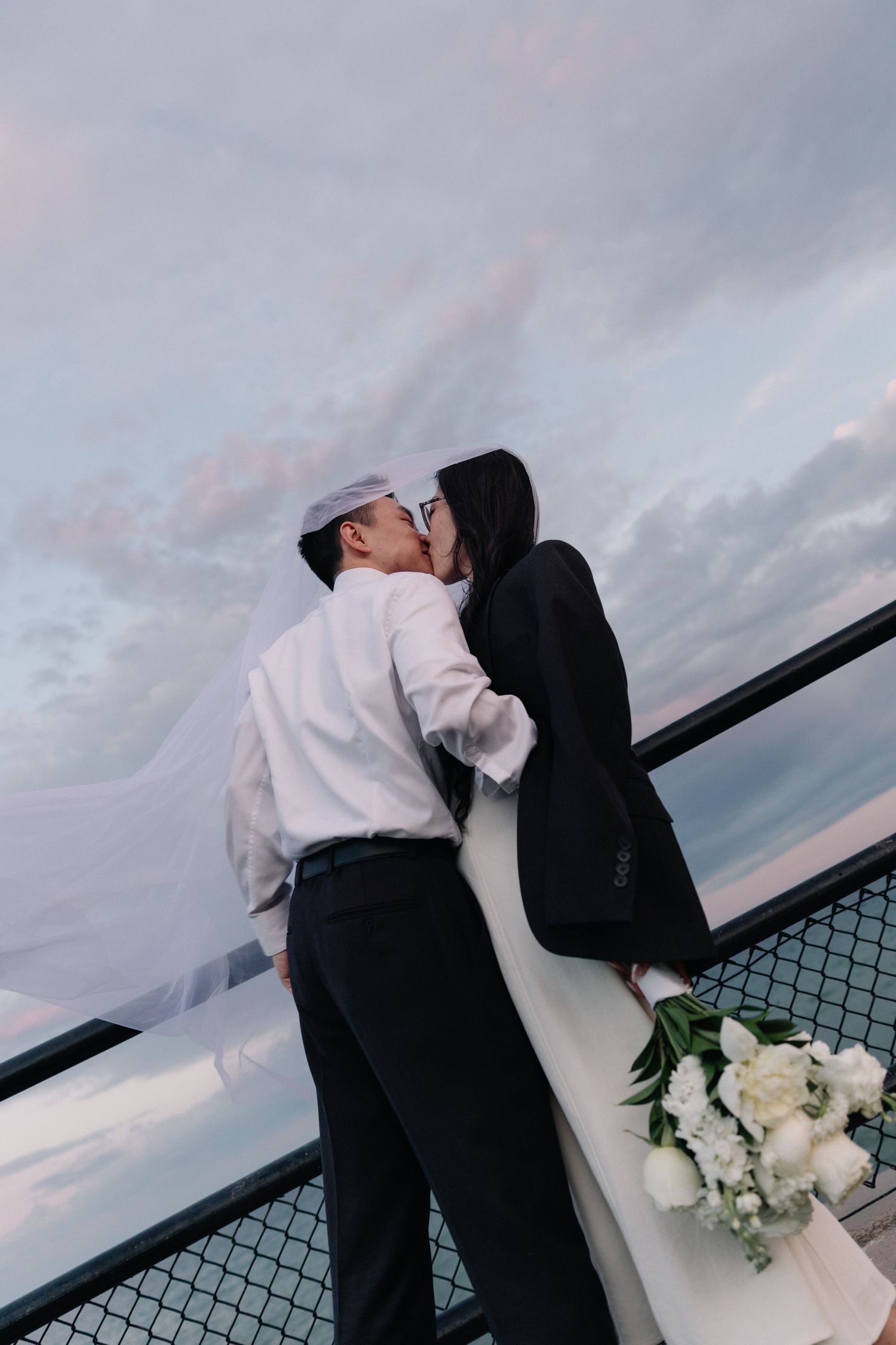A couple shares a romantic kiss on a rooftop at dusk, with a white bridal bouquet and dramatic cloudy sky backdrop.