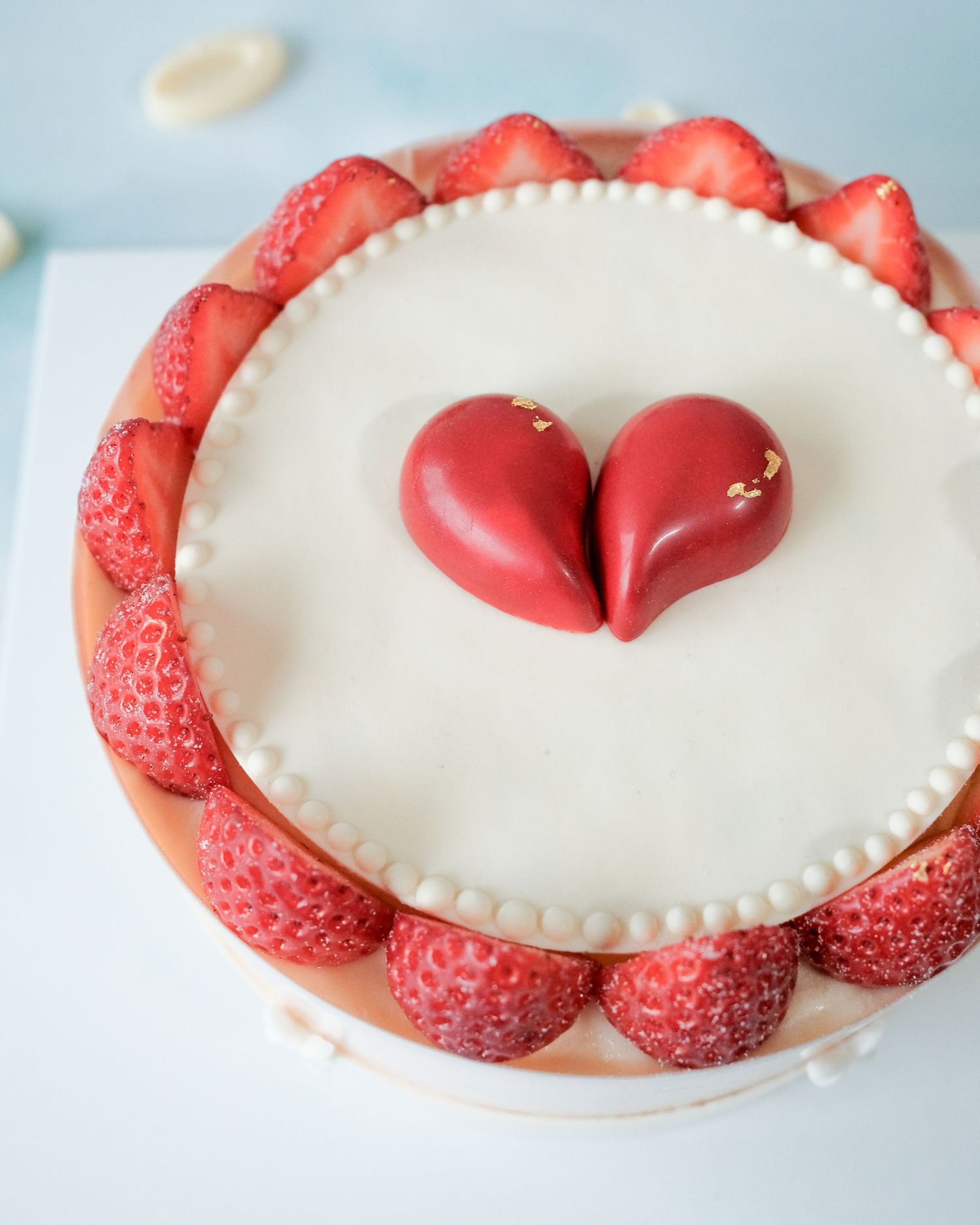 Elegant white cake with fresh strawberries around the edge and two red fondant hearts in the center.
