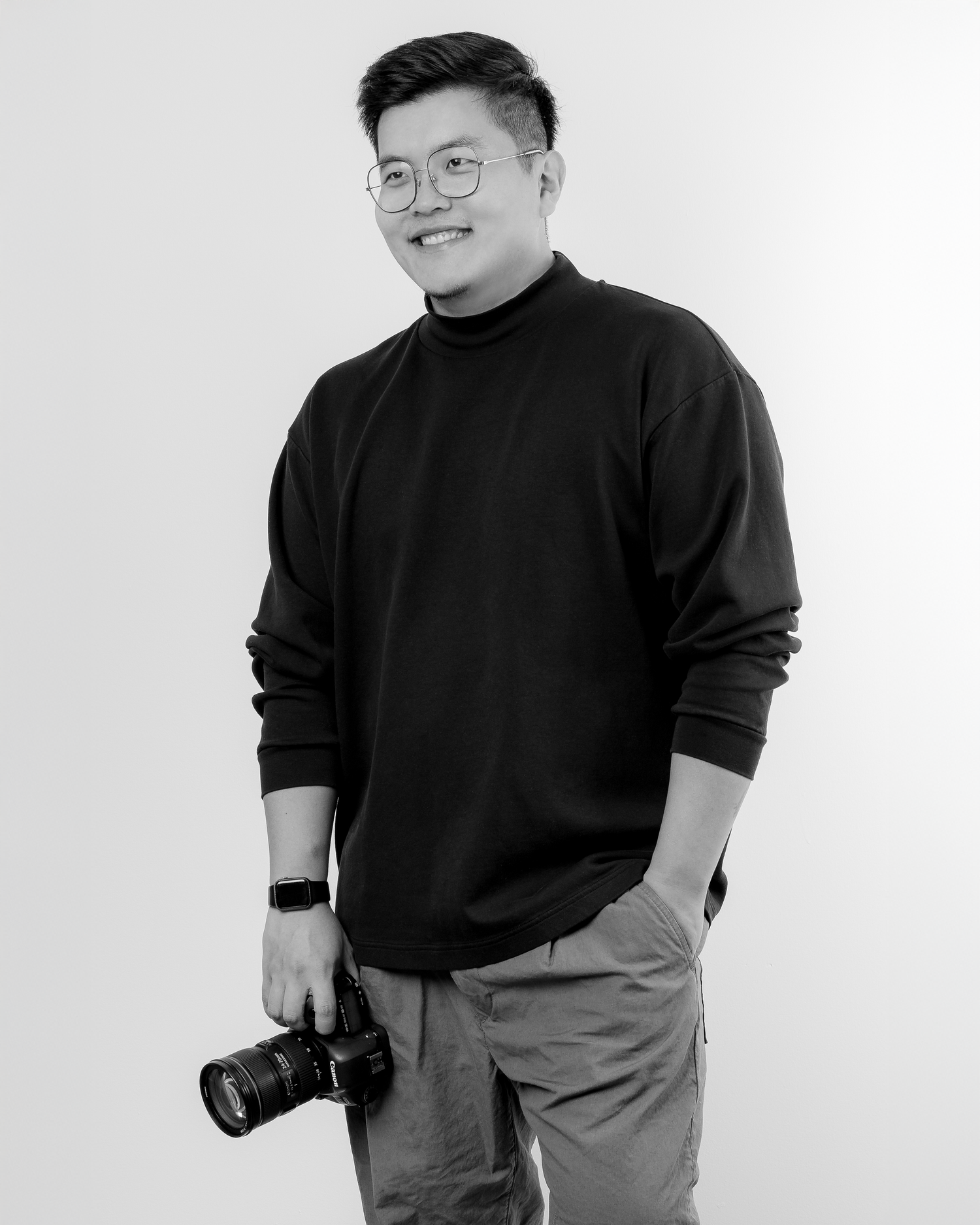 Portrait photographer in dark clothing poses with camera against white backdrop.
