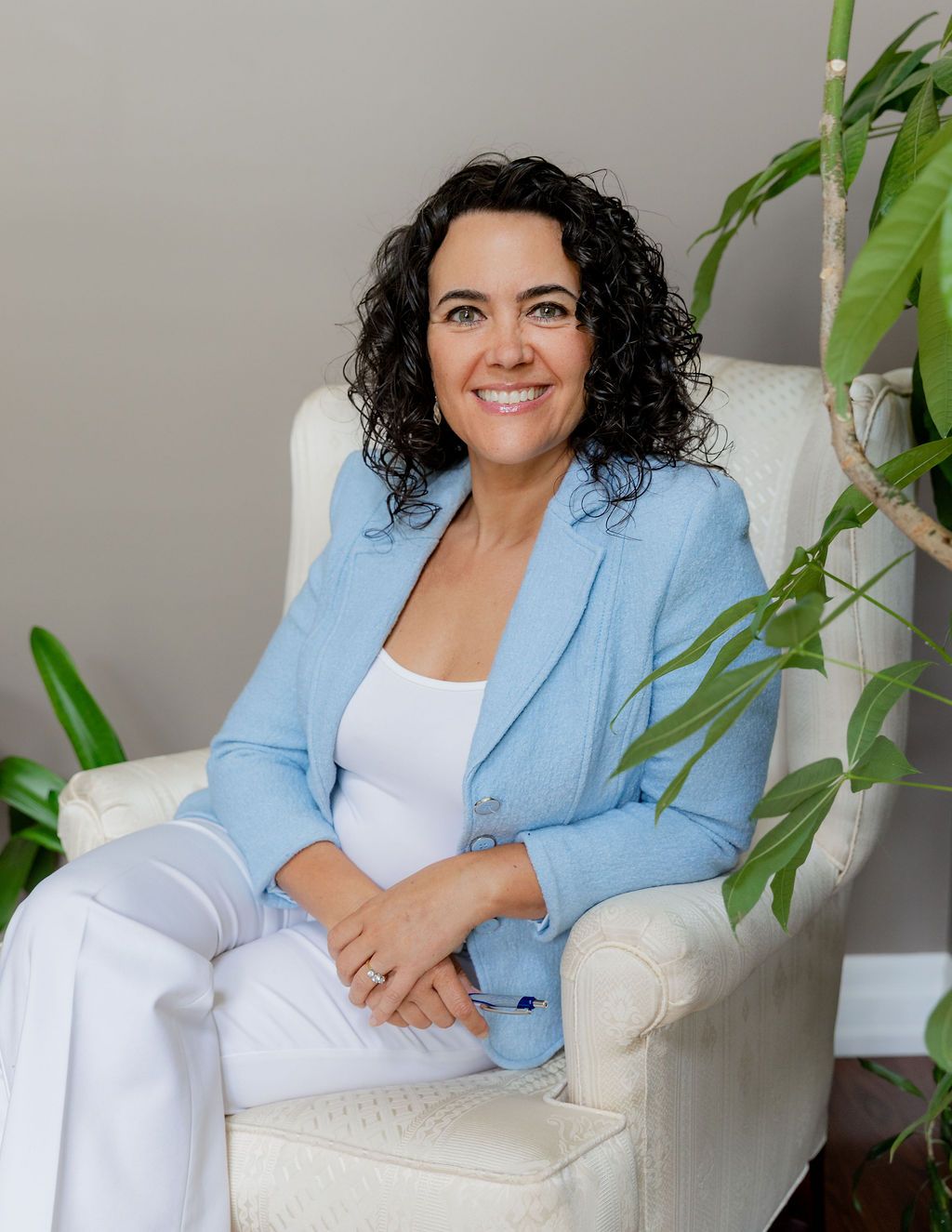 Professional woman in light blue blazer sitting in white armchair, smiling confidently in bright modern office setting.