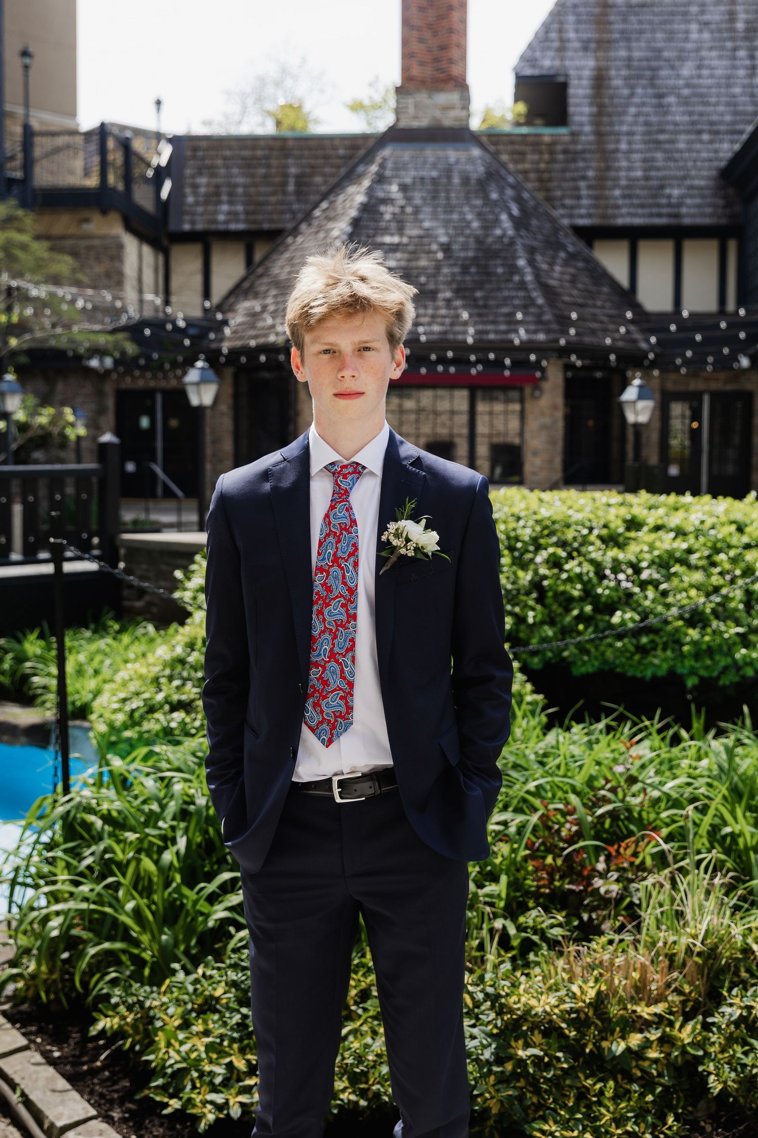 Young man in navy suit with red paisley tie and boutonniere stands outdoors before Tudor-style building.