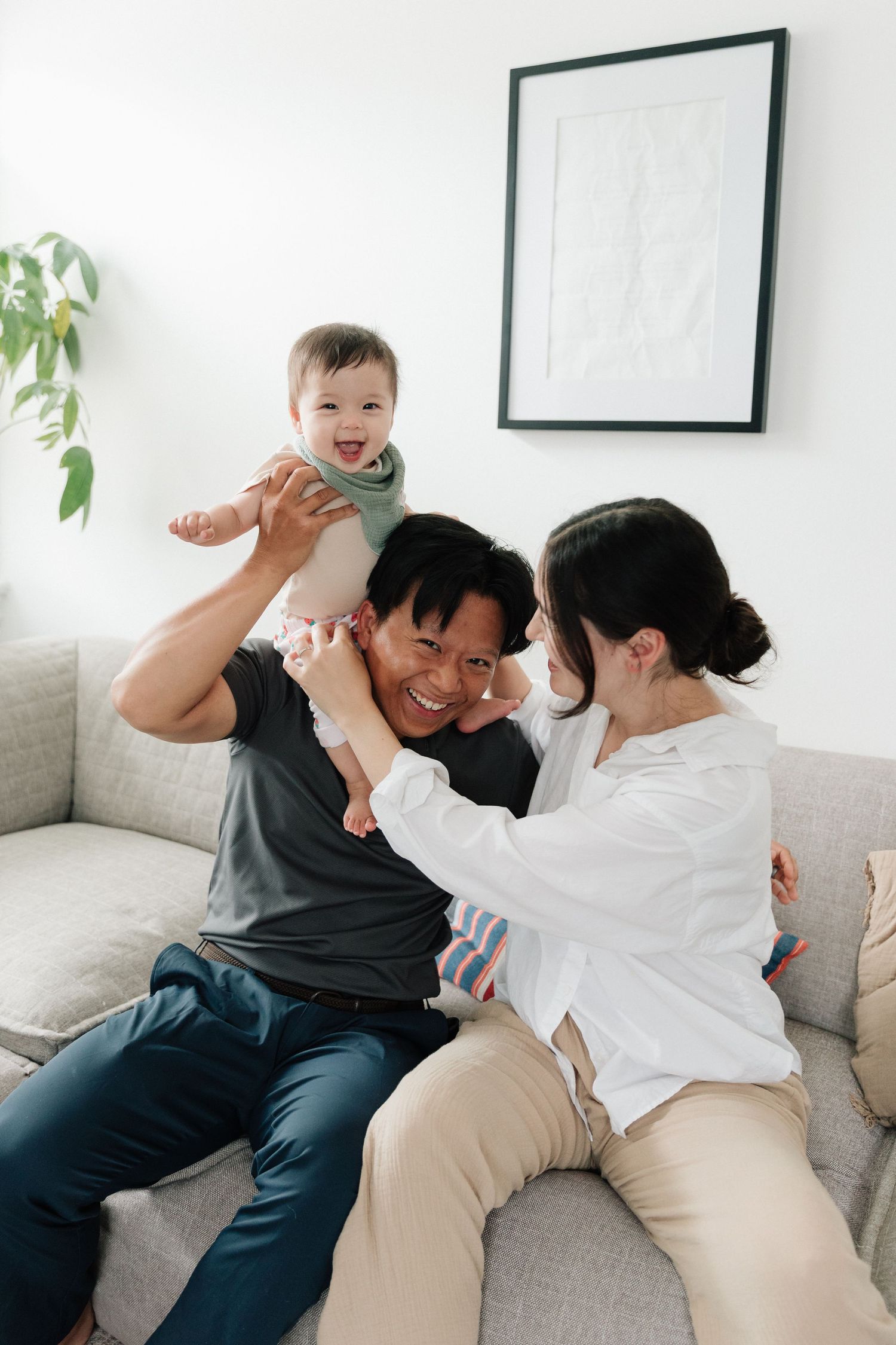 Happy Asian family with parents sitting on sofa lifting smiling baby in bright modern living room.