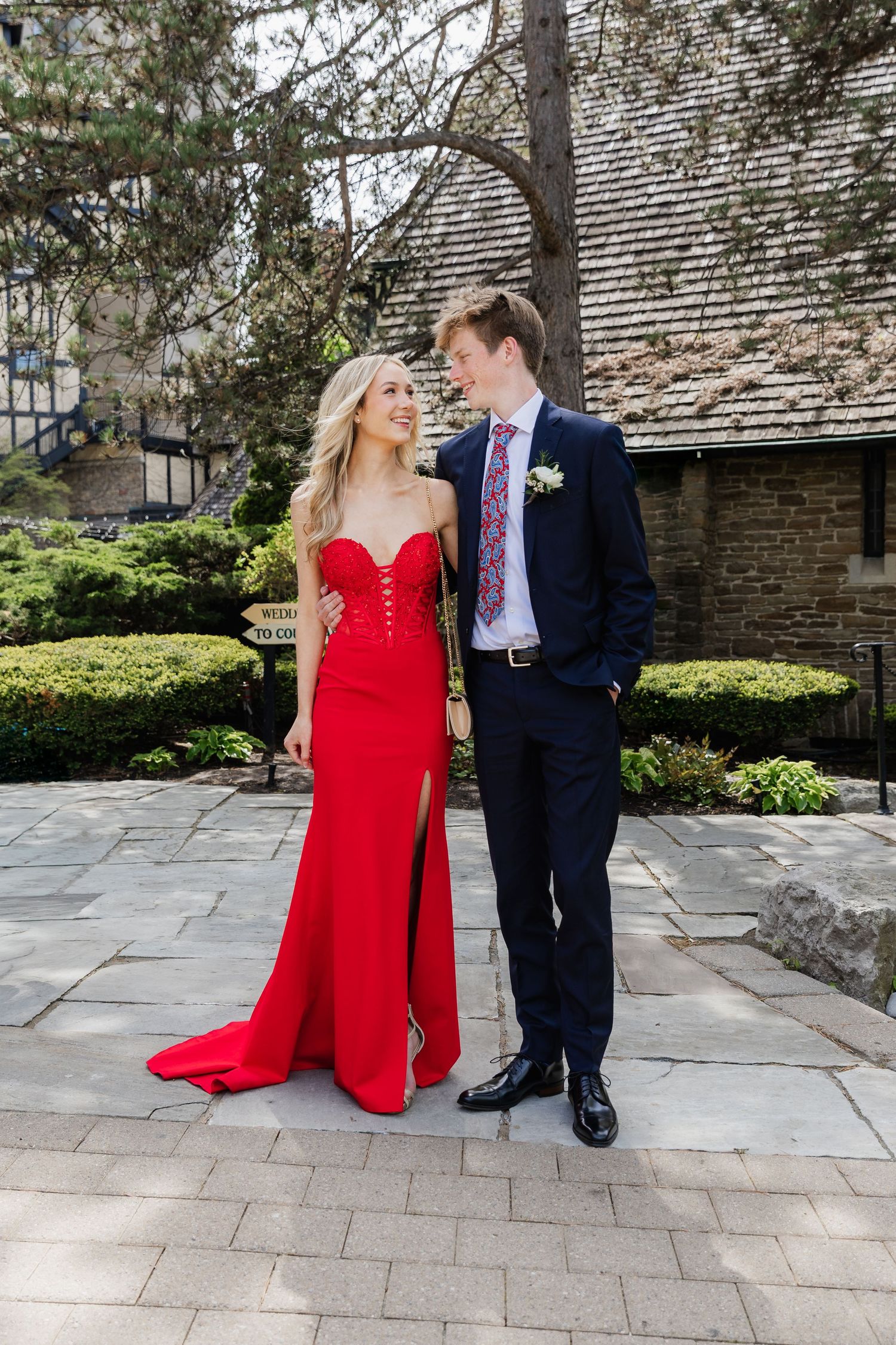 A young couple in formal attire, woman in red gown and man in navy suit, posing outdoors at a prom event.