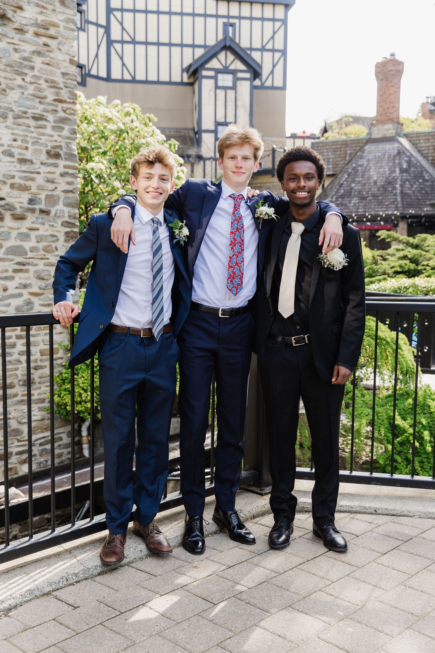 Three young men in suits and ties pose together on an outdoor balcony at a formal event.