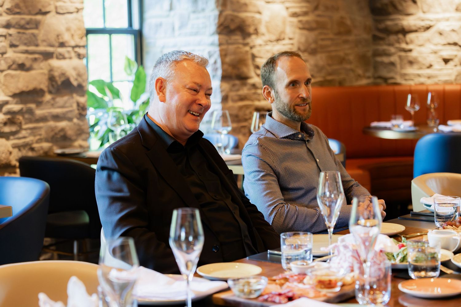 Two men smiling and laughing at a restaurant table with wine glasses and plates in a stone-walled dining room.