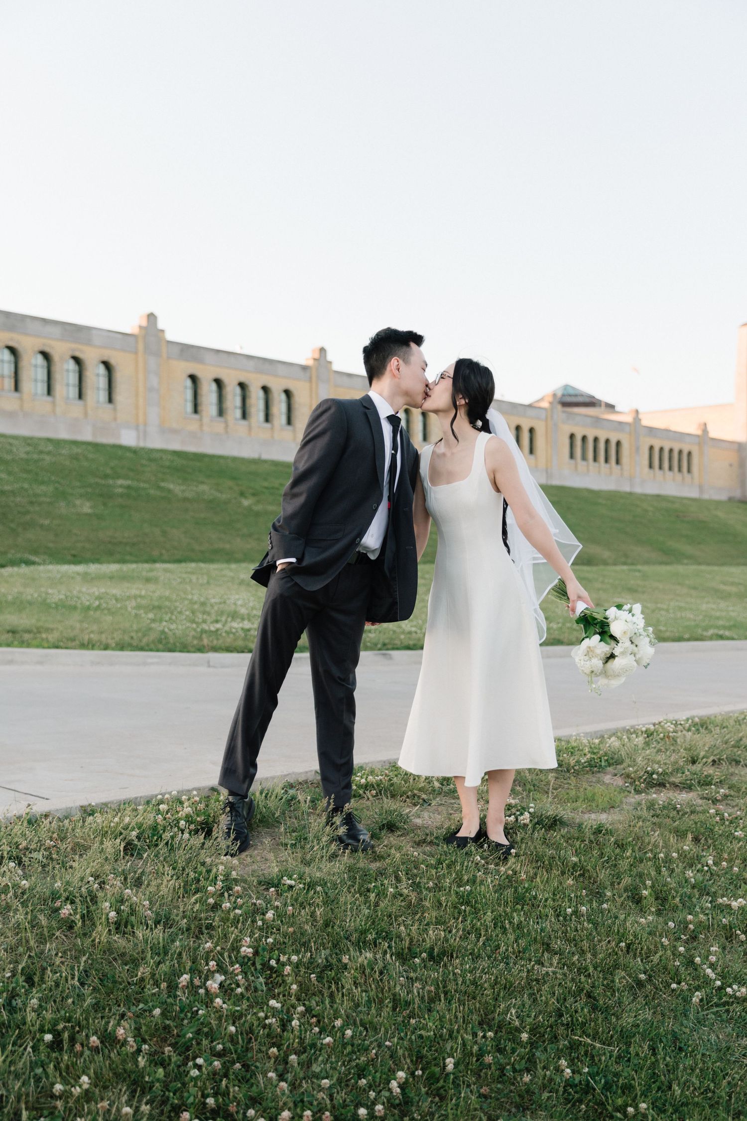 A bride and groom share a kiss outdoors on a grassy hill, she holds white flowers wearing a midi dress and veil.
