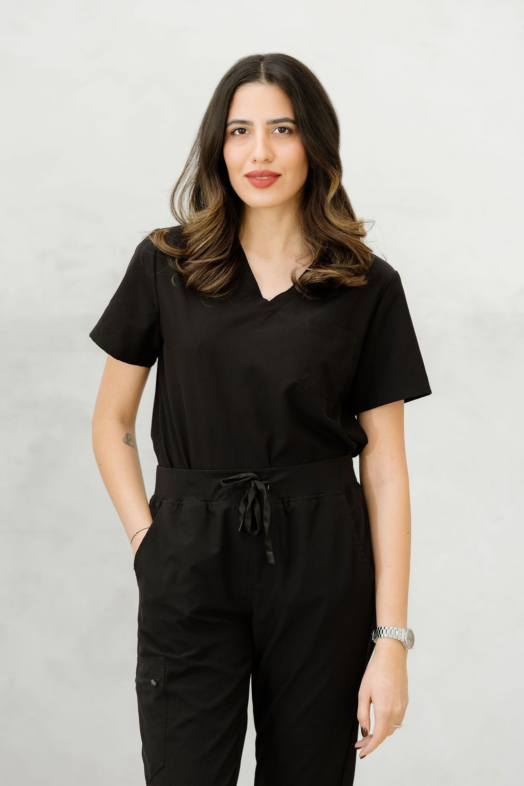 Woman in black medical scrubs set with drawstring pants and V-neck top, posing against light gray background.
