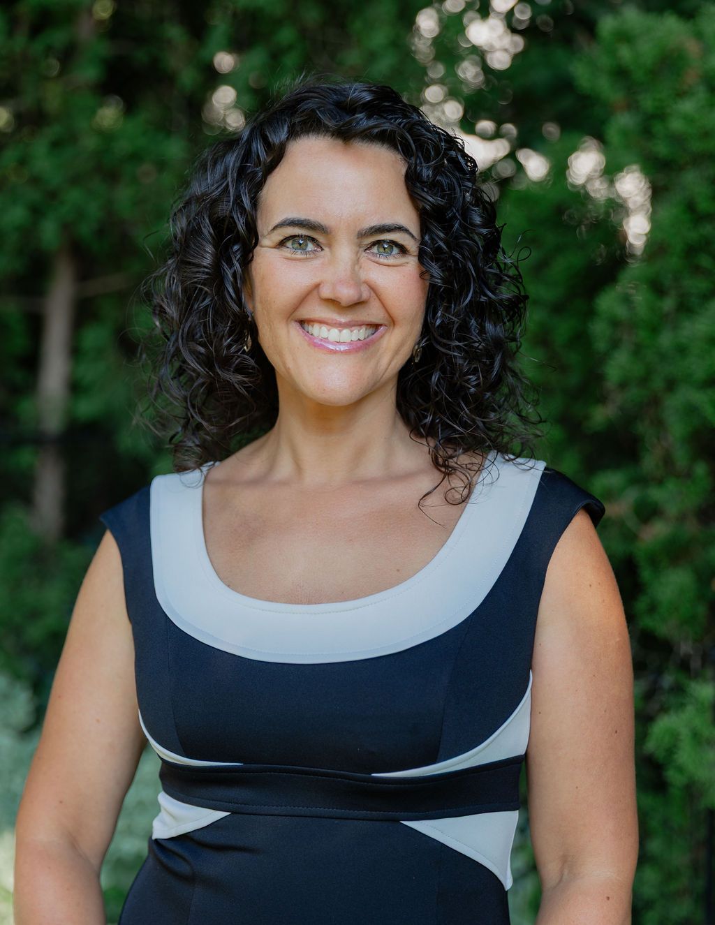 Smiling woman with curly dark hair wearing navy and gray sleeveless dress poses outdoors among green trees.