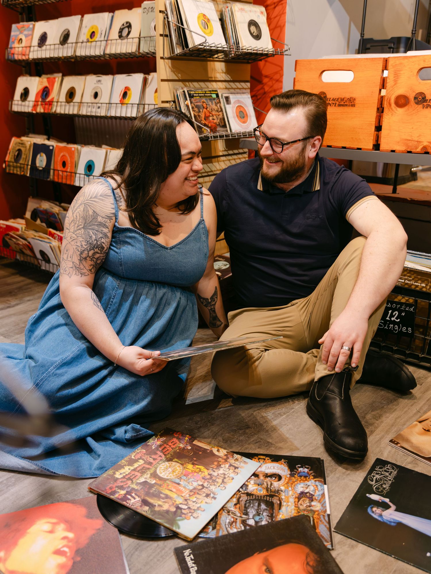 Couple sitting on floor of vinyl record shop, smiling at each other, surrounded by album covers and record bins.