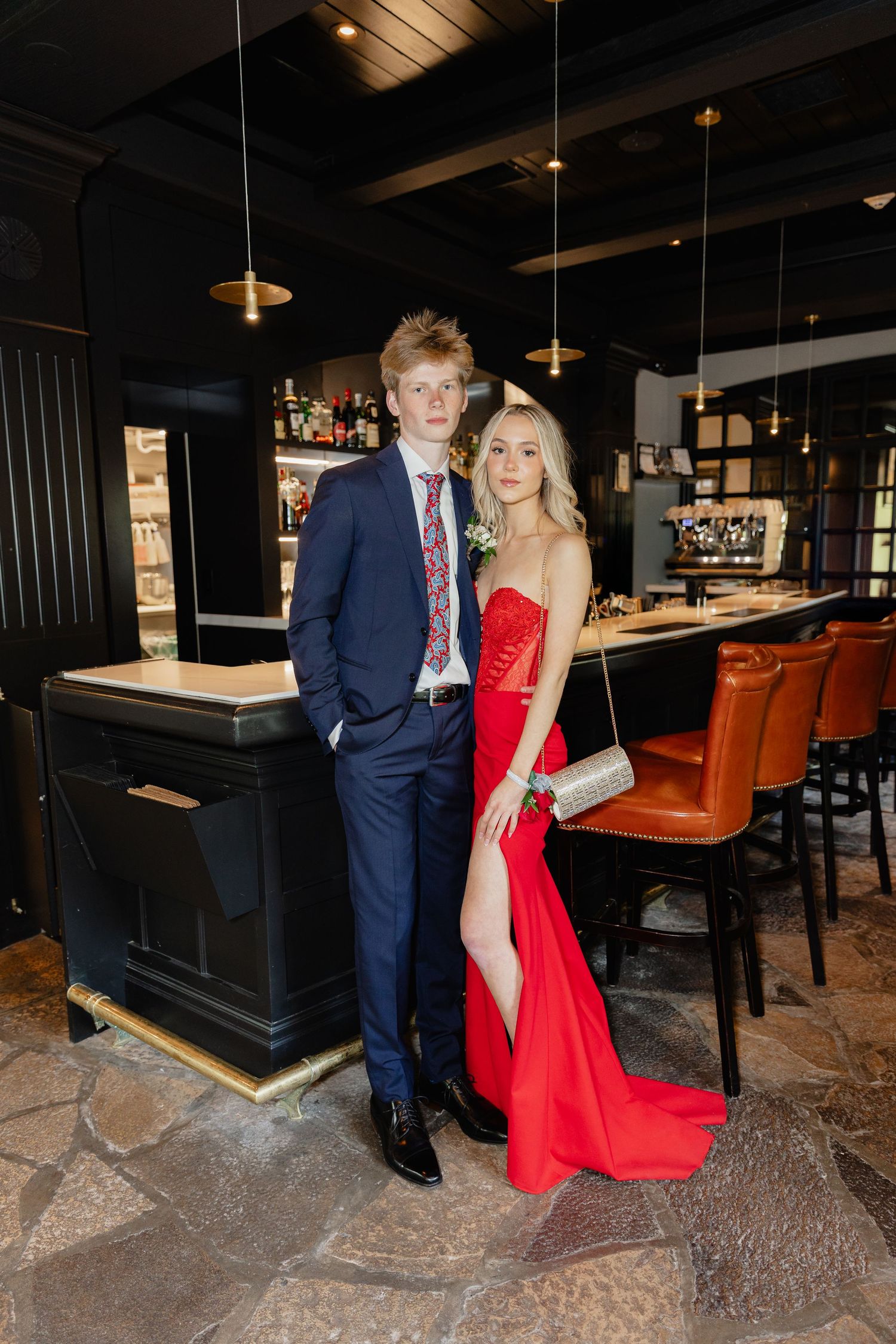 A young couple dressed in prom attire, him in a navy suit and her in a red gown, pose in an upscale restaurant bar.