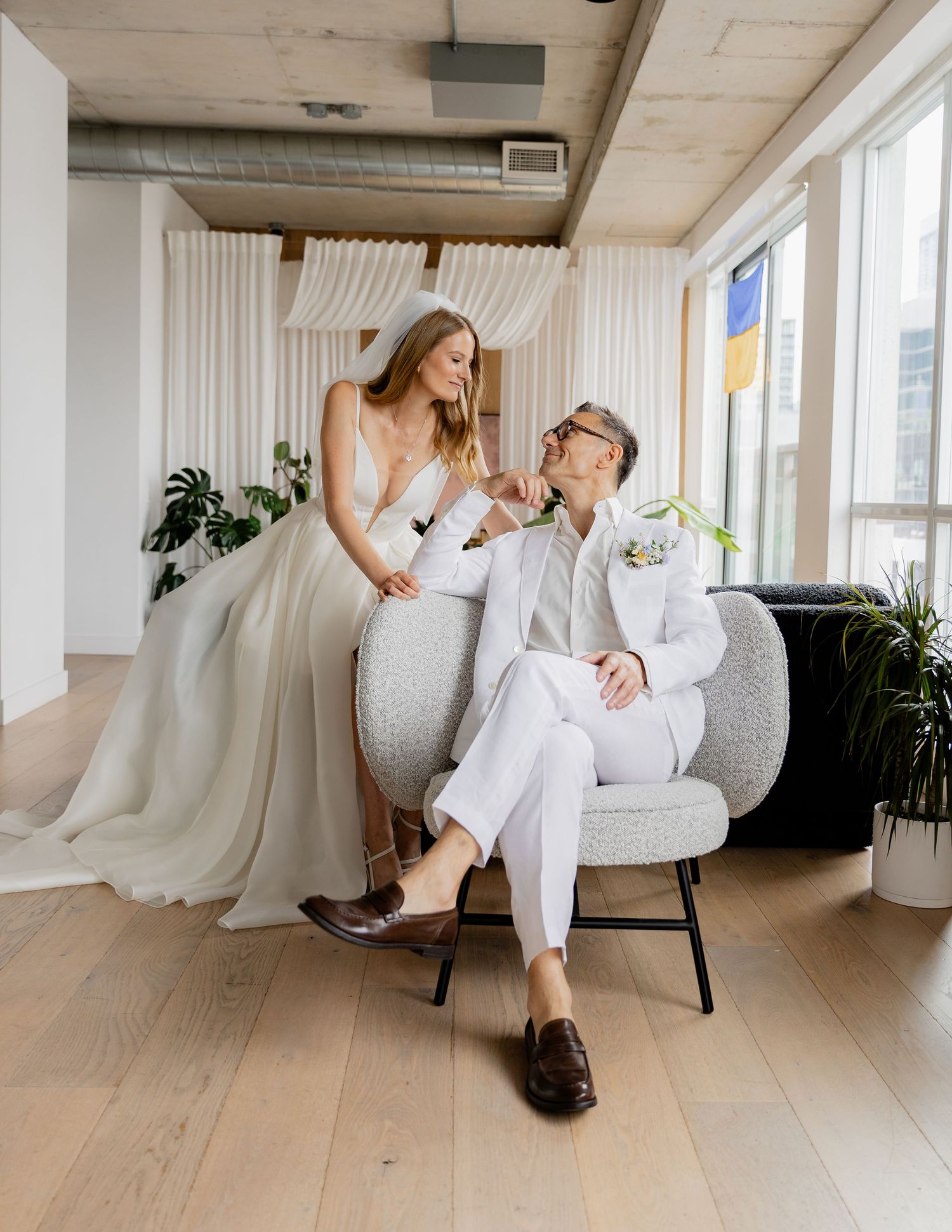 A couple in wedding attire shares a playful moment while sitting on a modern grey chair in a bright indoor space.