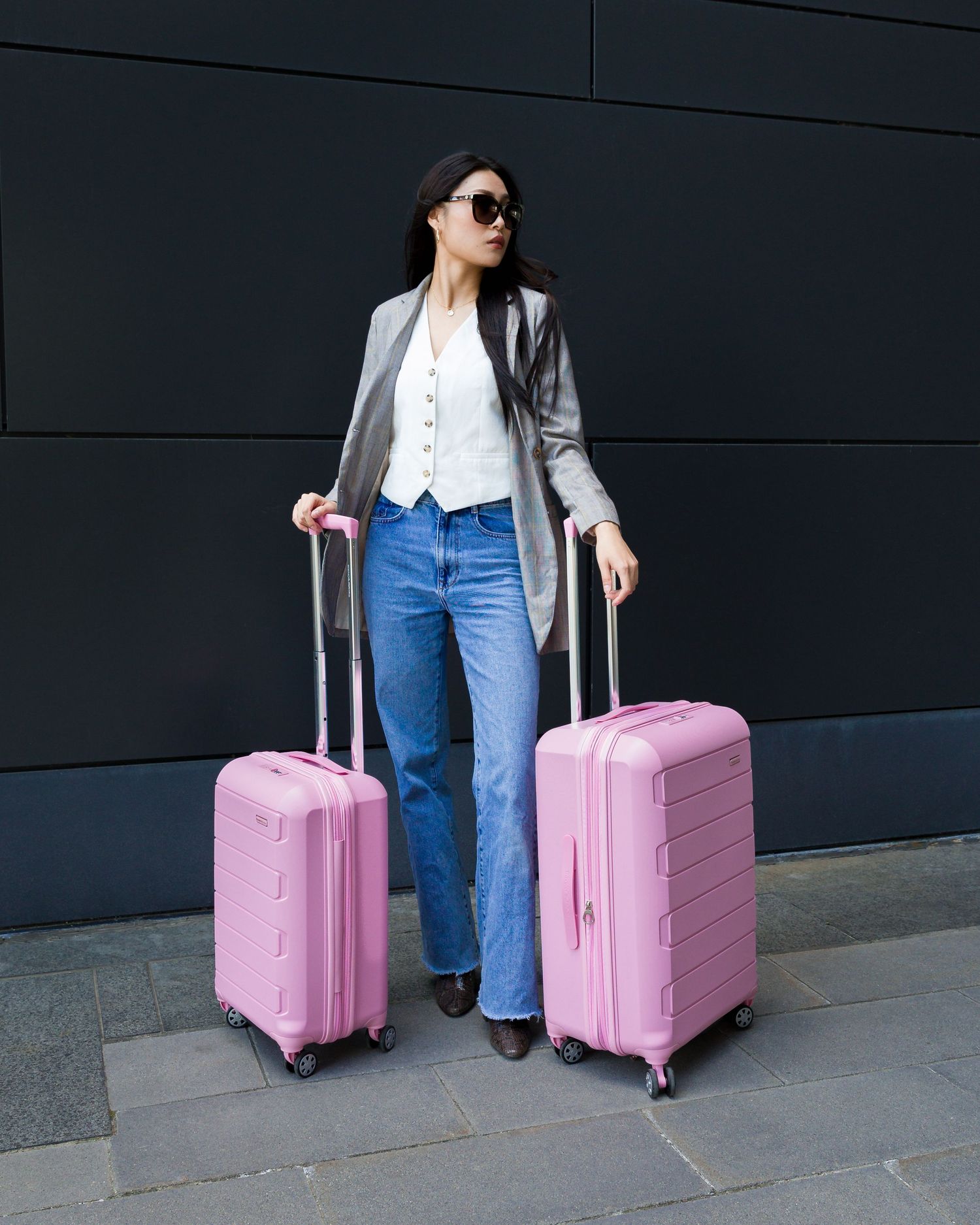 Casual outfit with grey blazer and jeans paired with pink rolling suitcase against dark wall.