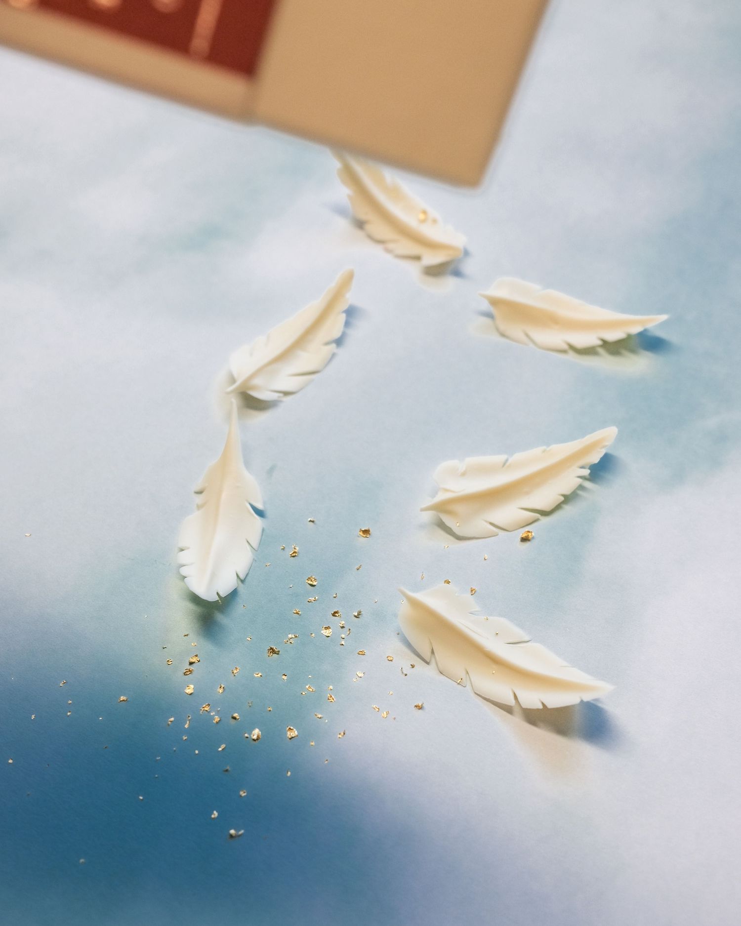 Delicate white feathers and gold glitter falling through a soft blue atmosphere.