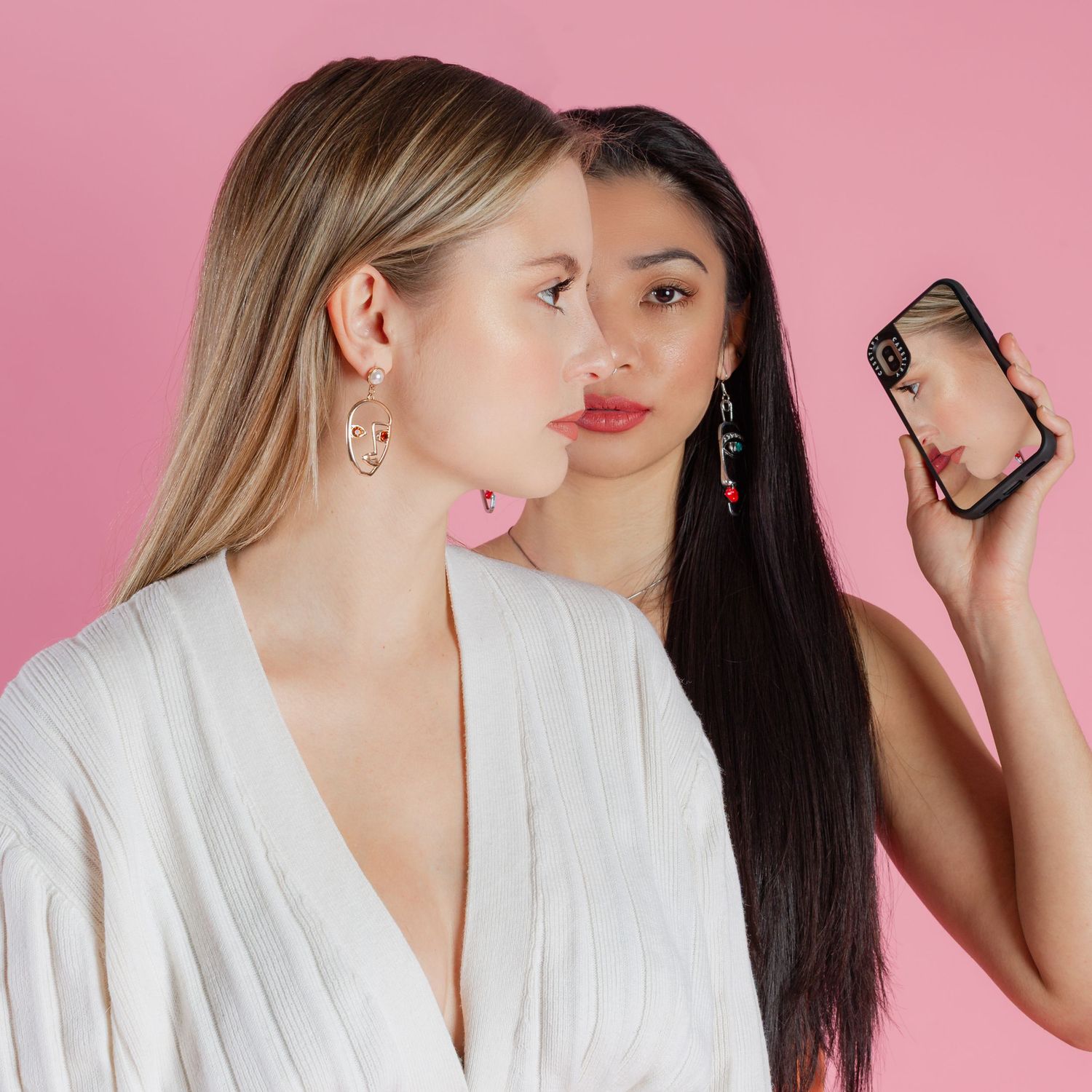 Two women in white robes posing with phones against pink background.