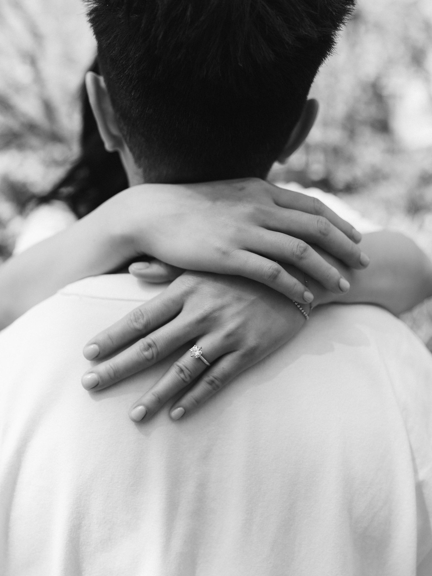 Black and white close-up of a woman's hands with rings wrapped around a man's shoulders in an embrace.