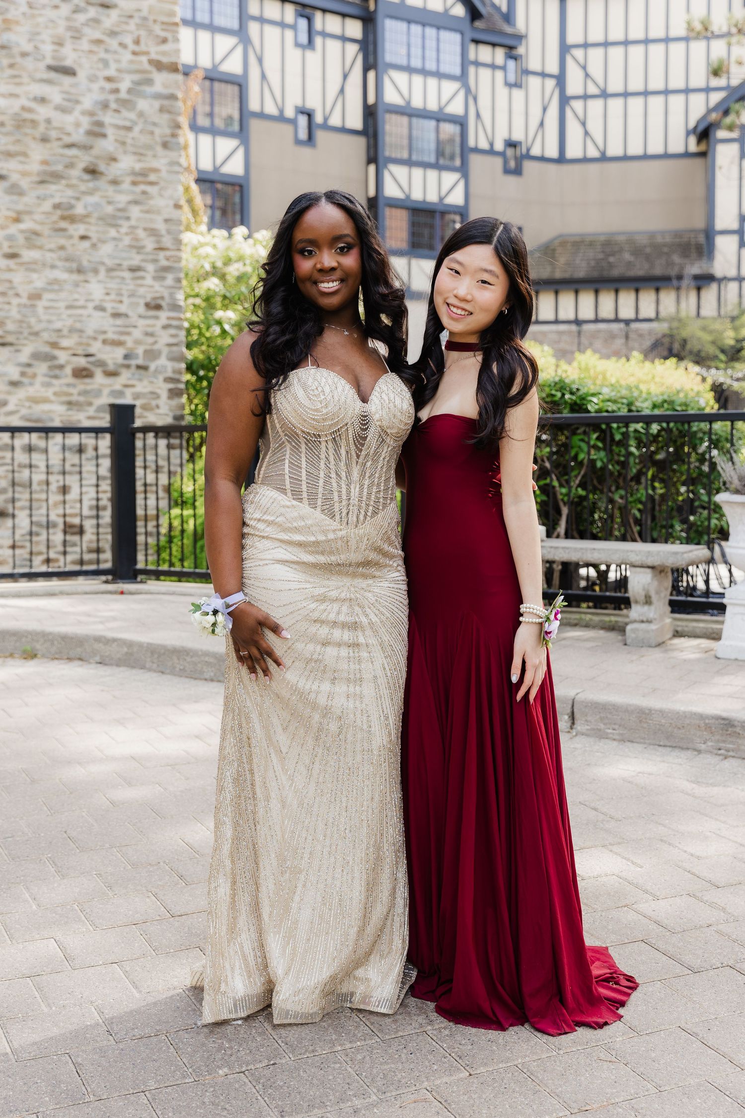 Two young women in elegant prom gowns, one gold beaded and one burgundy, posing outdoors at a formal event.
