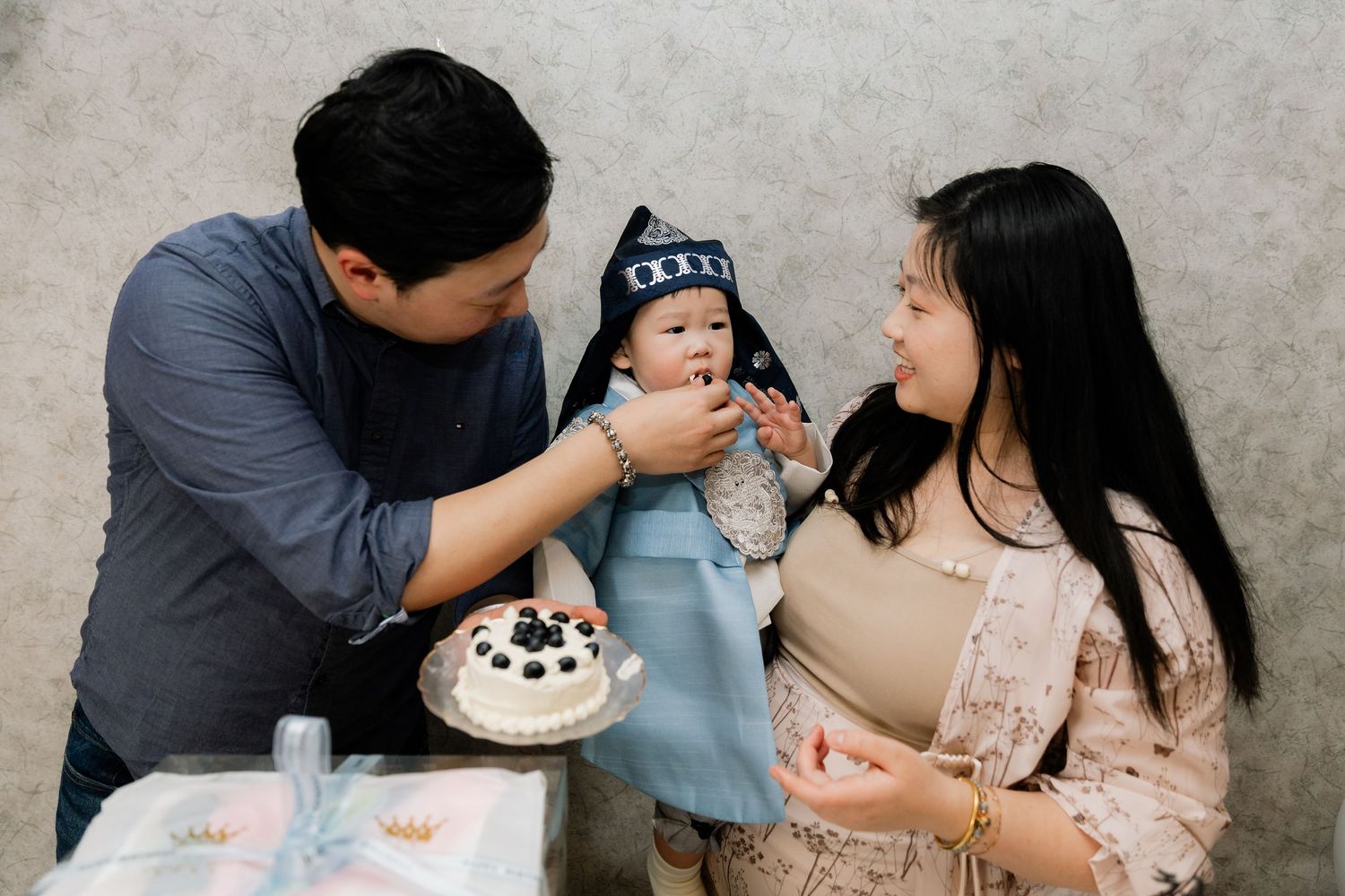 Asian family celebrating baby's first birthday with a white cake, parents smiling at infant in traditional outfit.