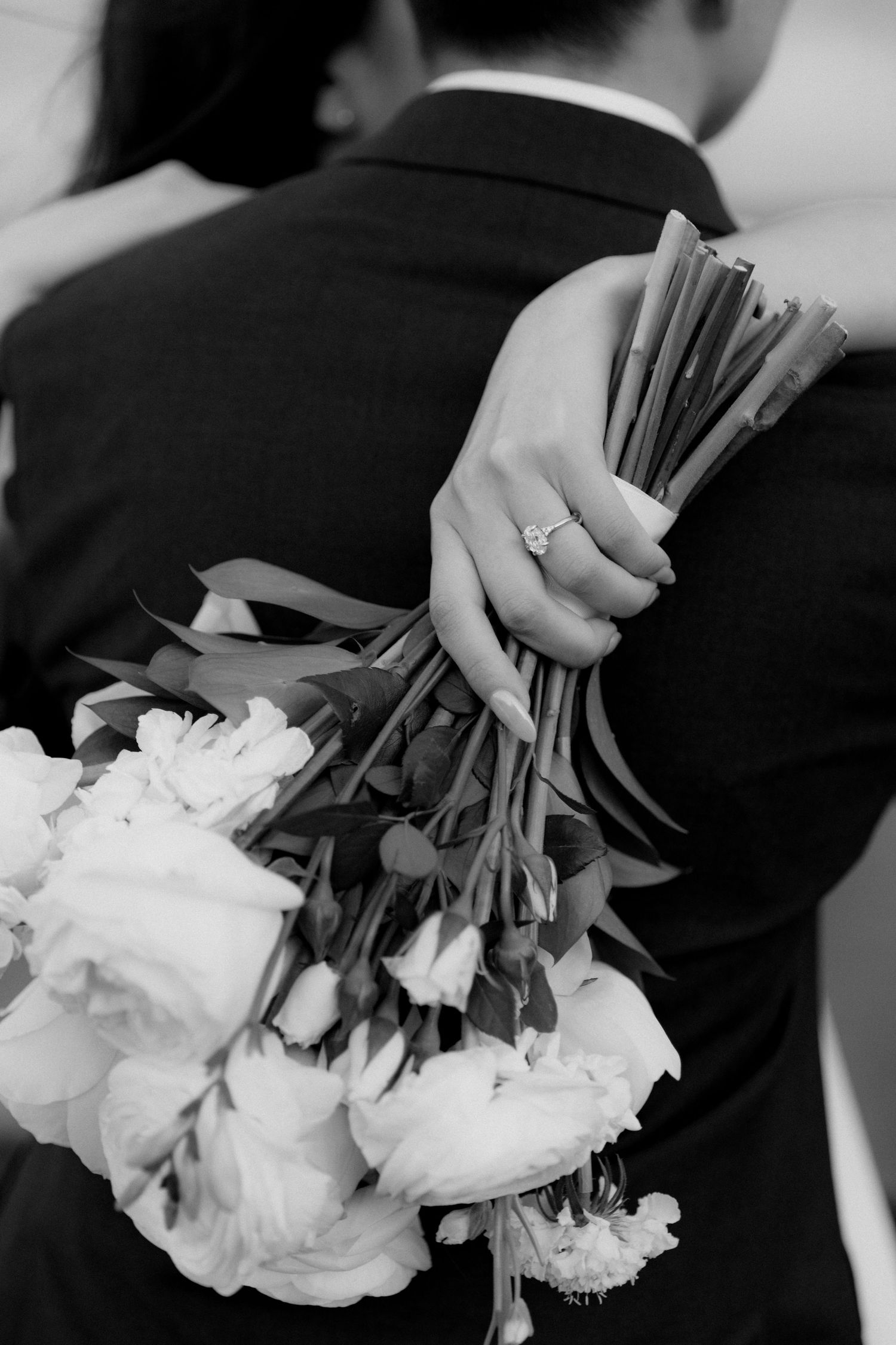 Black and white photo of a bride holding a floral bouquet while embracing a groom in a suit, engagement ring visible.