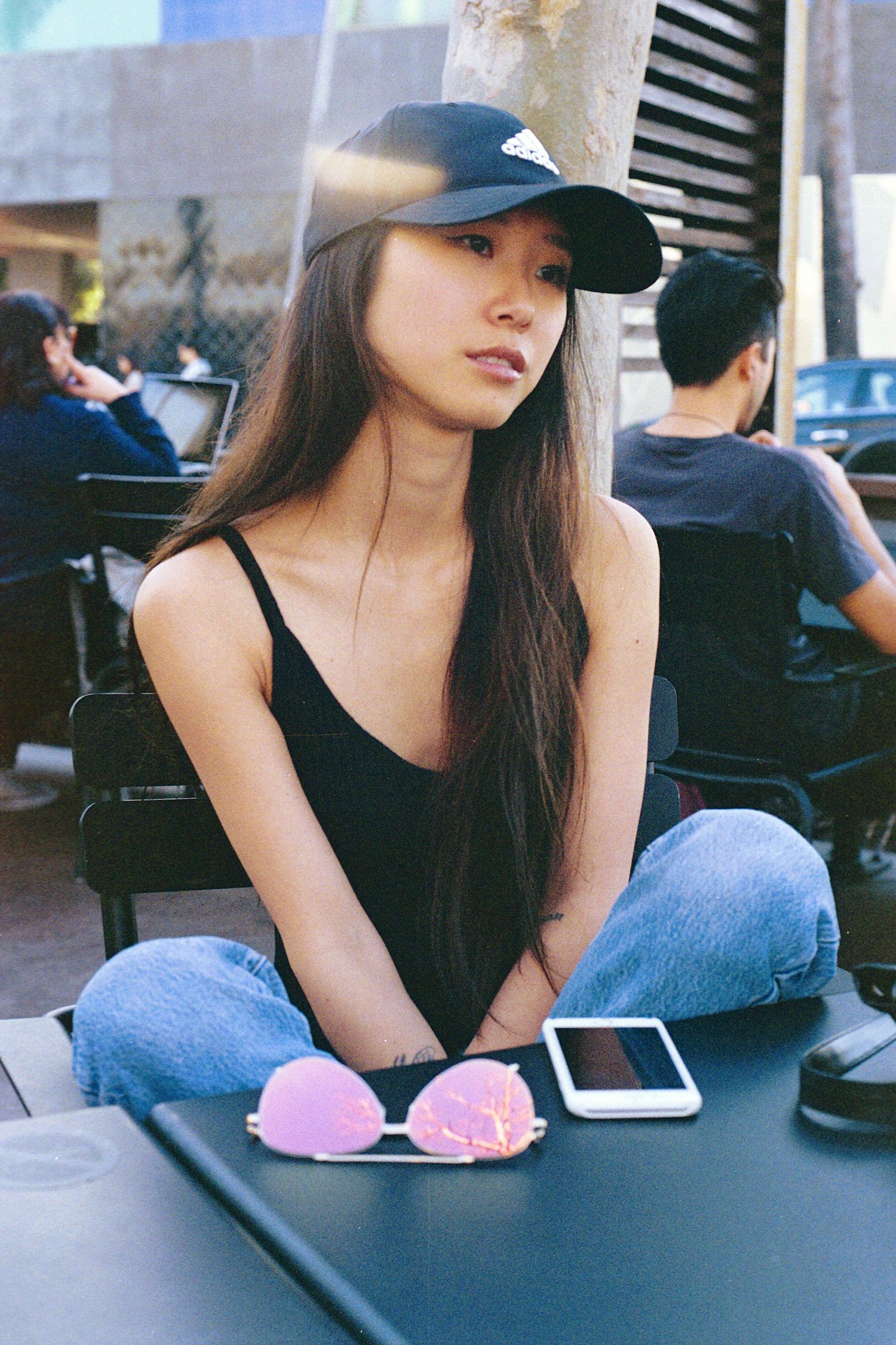 Person in black tank top and jeans sitting at outdoor table with phone.