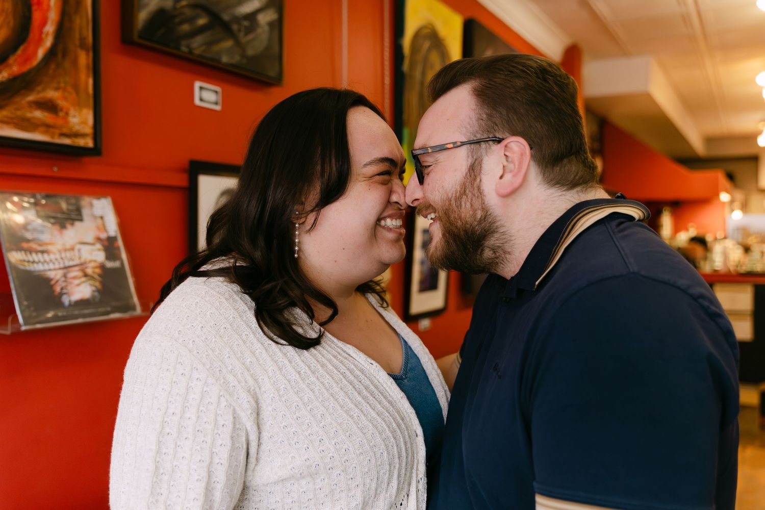 A couple shares a tender nose-to-nose moment, smiling intimately in a colorful restaurant with framed artwork on red walls.