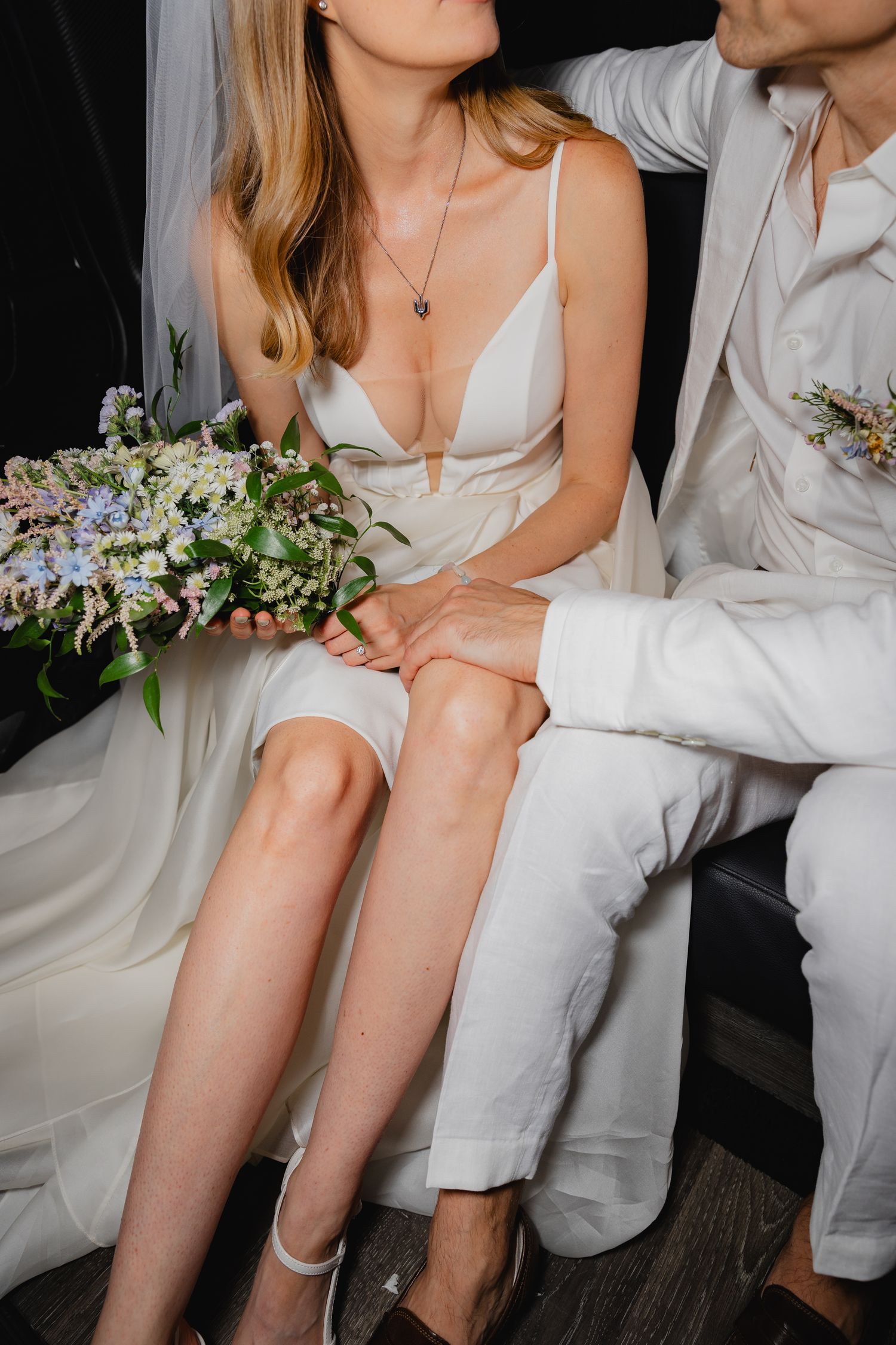 Couple in white wedding attire sitting close together with floral bouquet.