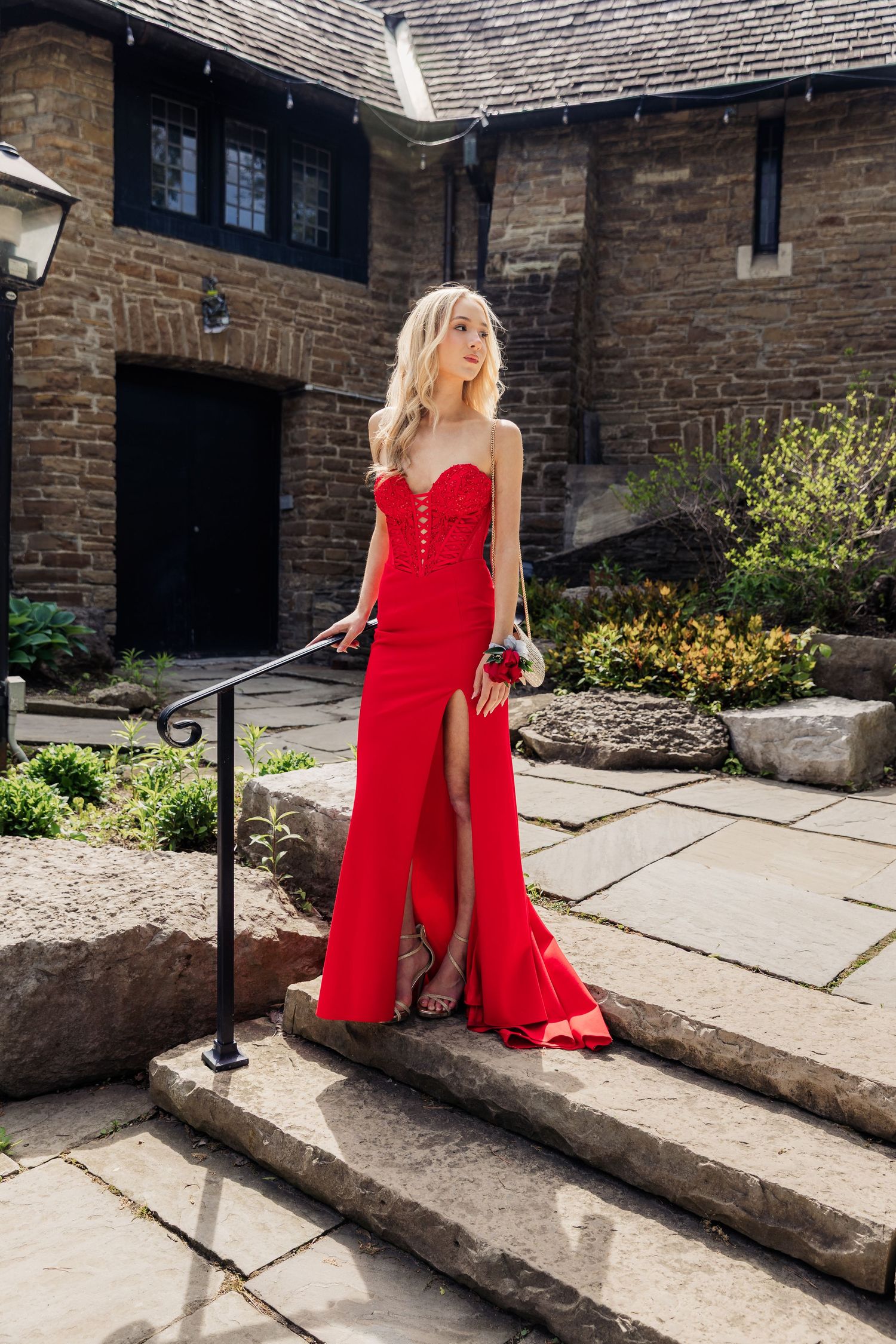 Blonde woman in elegant red strapless slit gown poses on stone steps outside a historic building.