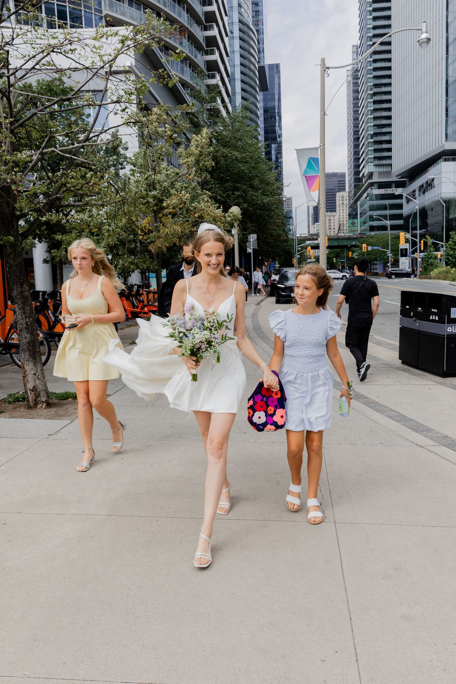 Three people in light-colored dresses walk down a city sidewalk with high-rise buildings in the background.