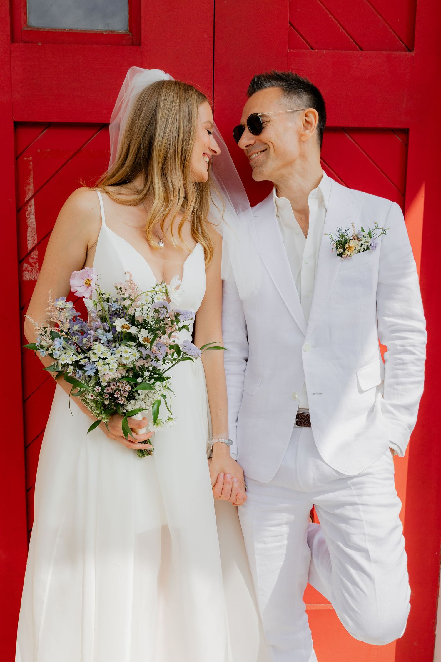 A couple in white wedding attire poses against a vibrant red wall with a delicate bouquet of flowers.