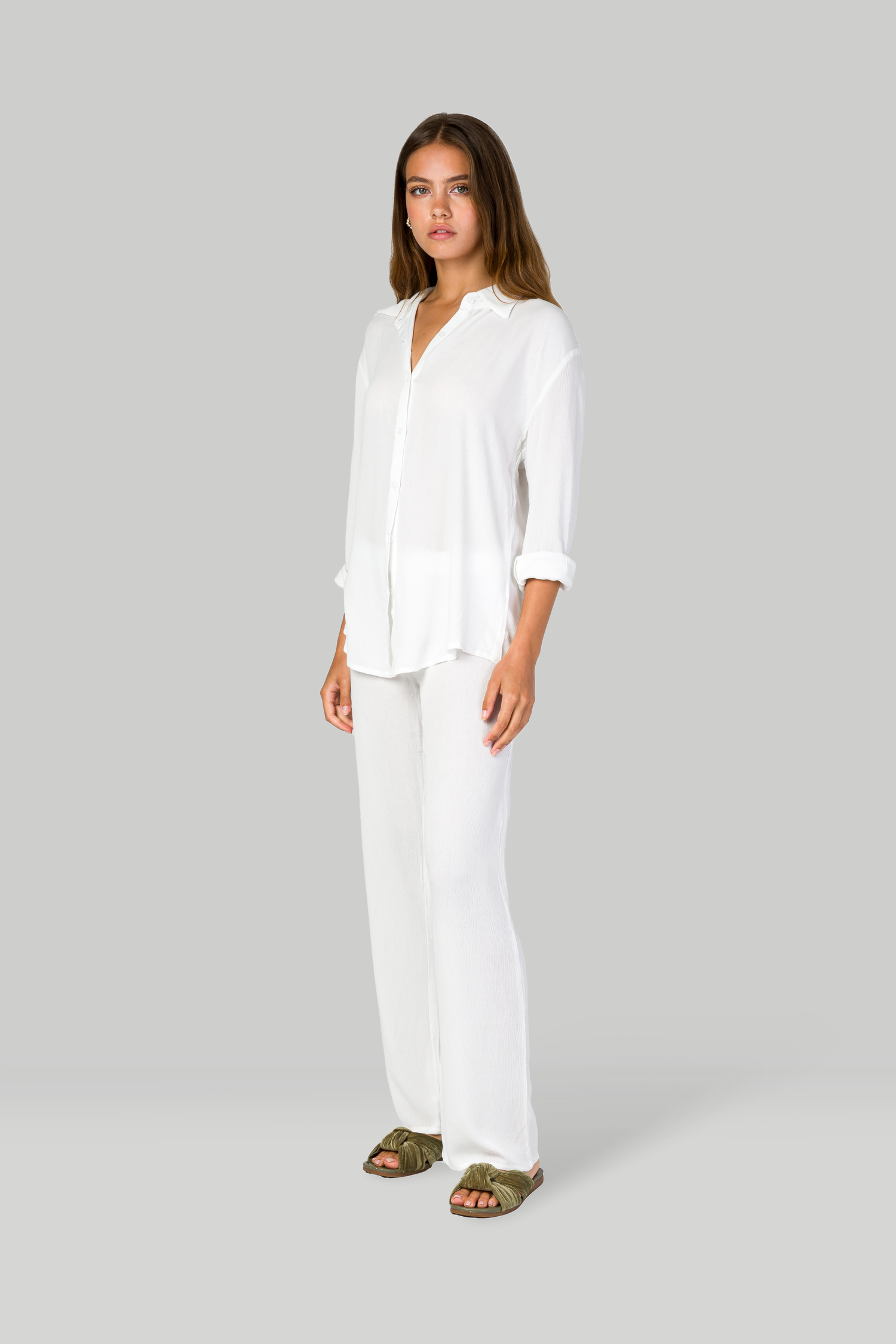 Model in all-white ensemble with flowing button-down shirt and relaxed pants against gray backdrop.