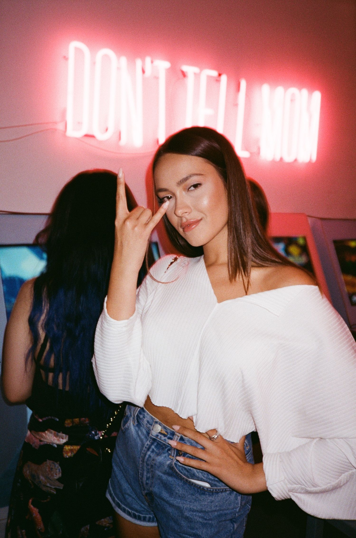 Person in white sweater and jeans posing under pink neon lights.