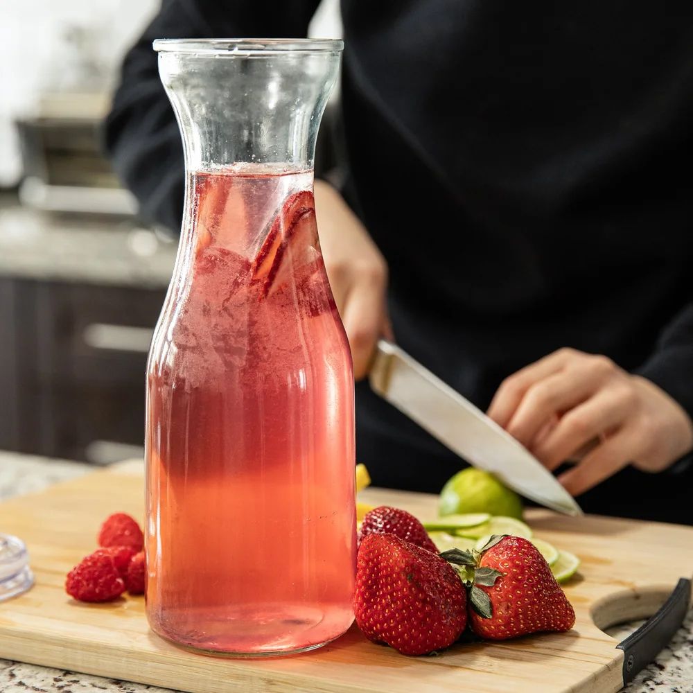 Fresh strawberries next to glass carafes filled with pink-colored beverage on a wooden cutting board.