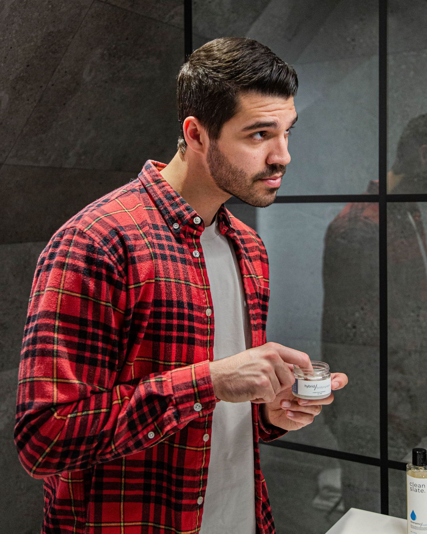 Person wearing red plaid shirt examining skincare product in bathroom.