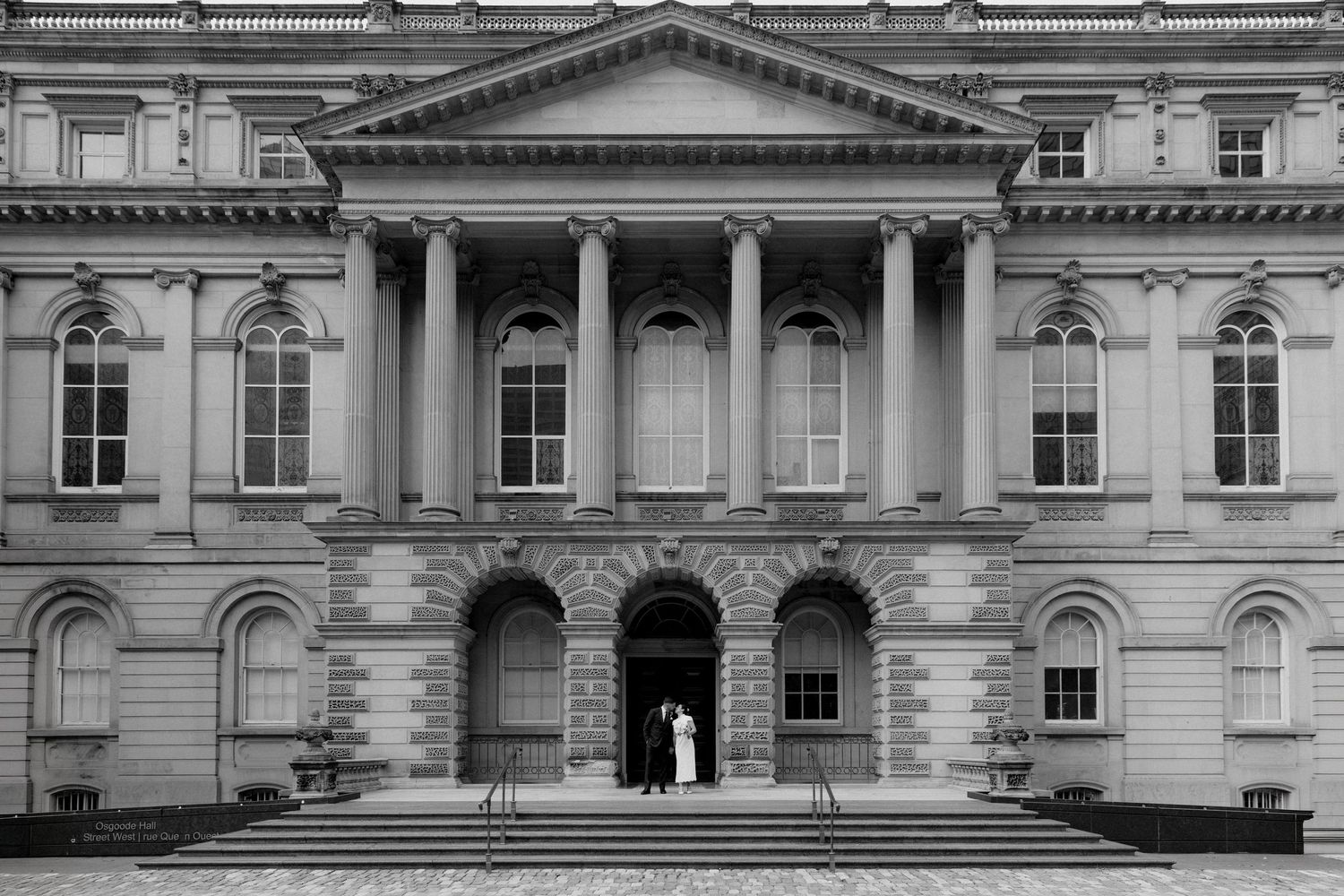 Black and white photo of a couple standing at the entrance of a grand neoclassical building with tall columns.