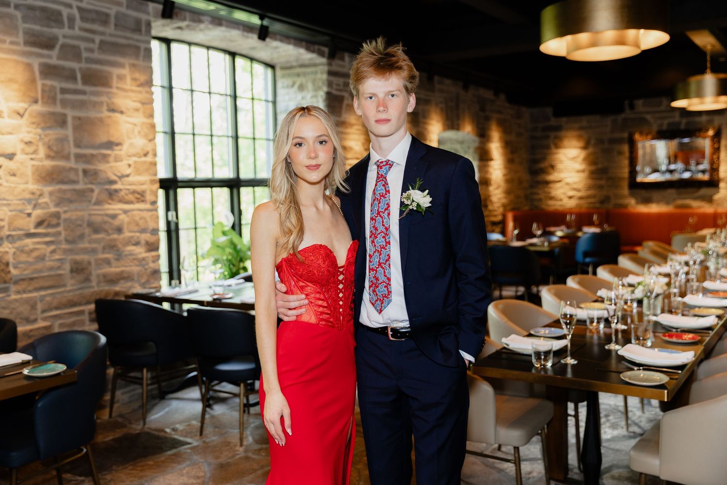 Young couple in formal attire posing in a restaurant, girl in red dress and boy in navy suit with floral tie.