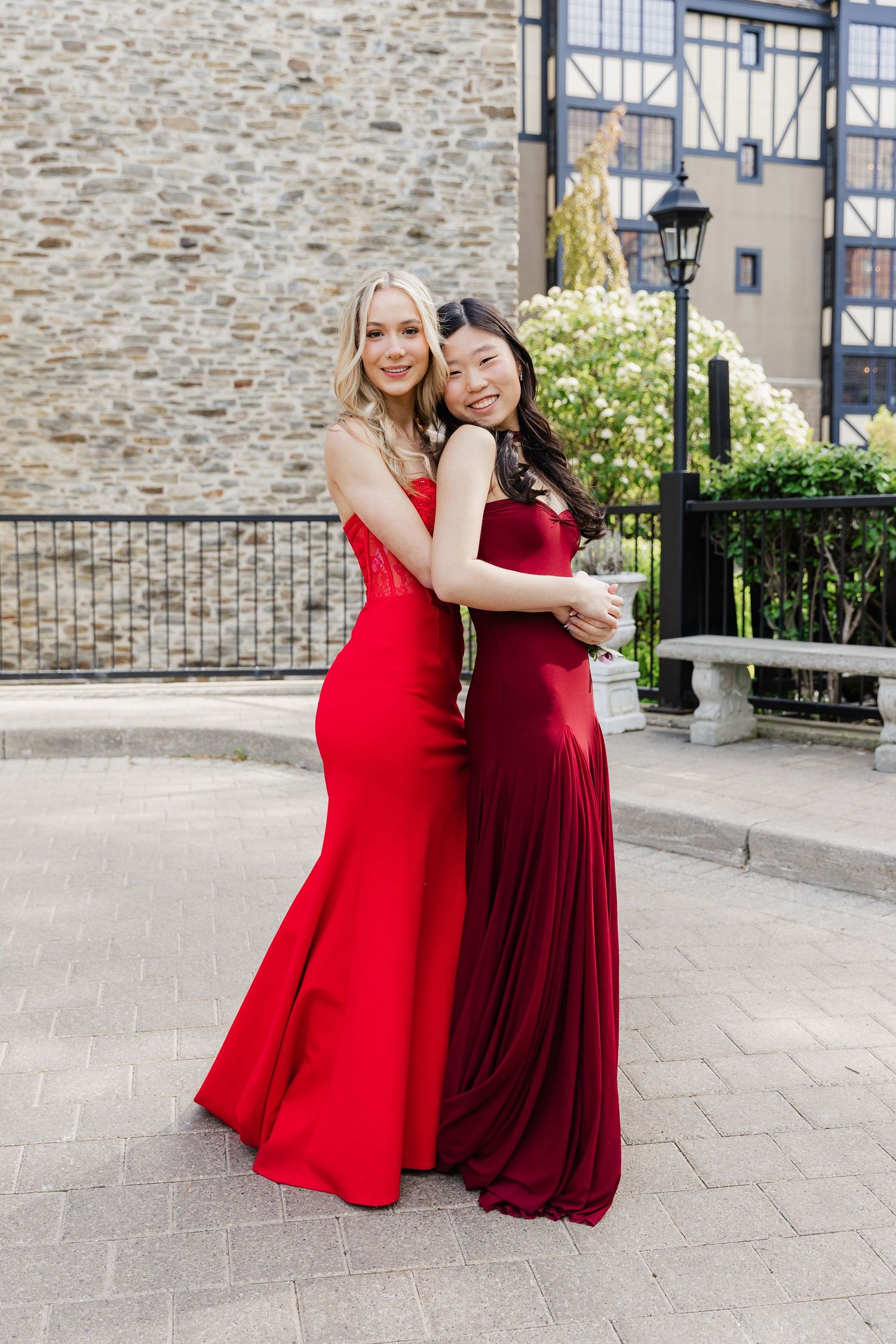 Two young women in matching red formal gowns embrace outdoors on a stone plaza, smiling for a prom photo.