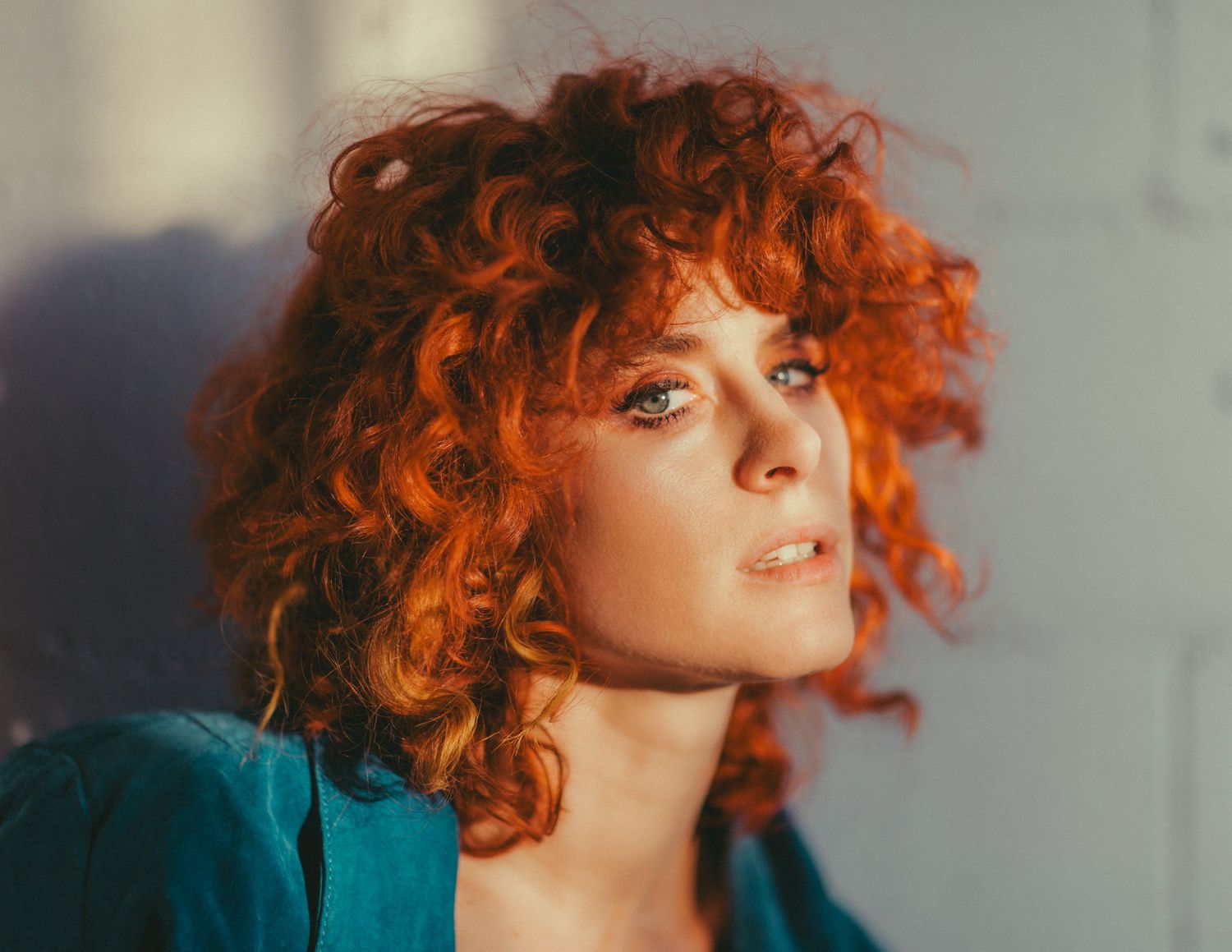 Vibrant red curly hair portrait with teal blazer in soft window light.