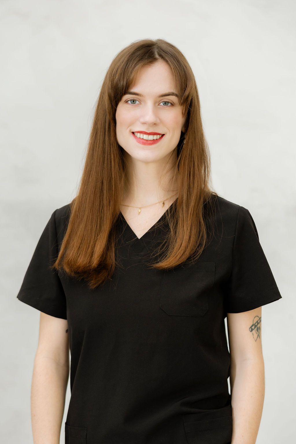 Smiling woman with long brown hair wearing a black v-neck scrub top and gold necklace, with a tattoo on her arm.