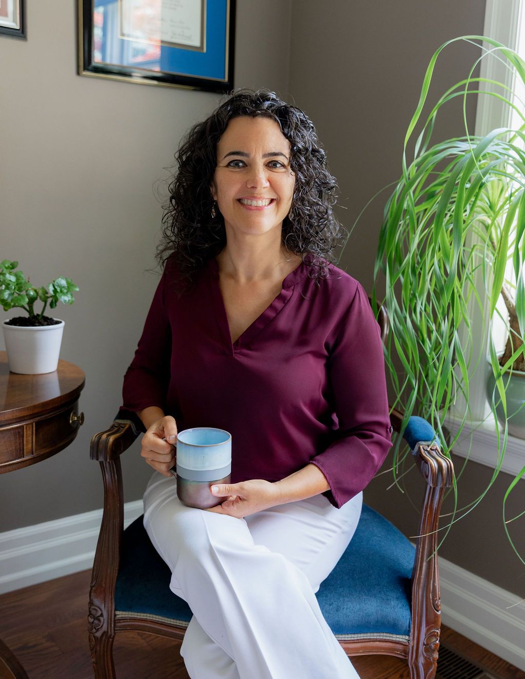 Woman with curly hair sitting in a chair, wearing a burgundy top and white pants, holding a mug.