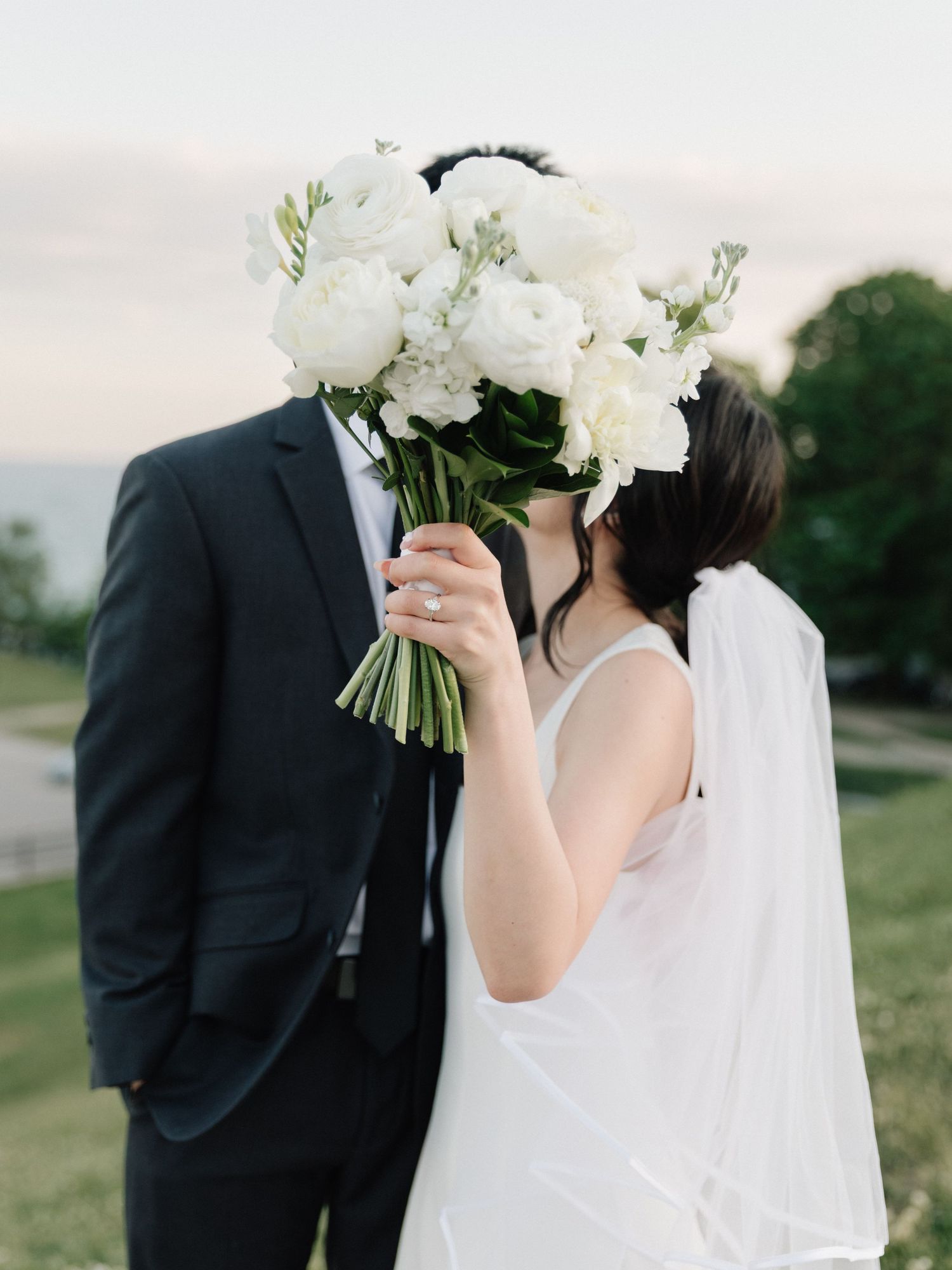 Bride and groom kissing behind a lush white peony bouquet outdoors, wearing wedding dress with veil and black suit.