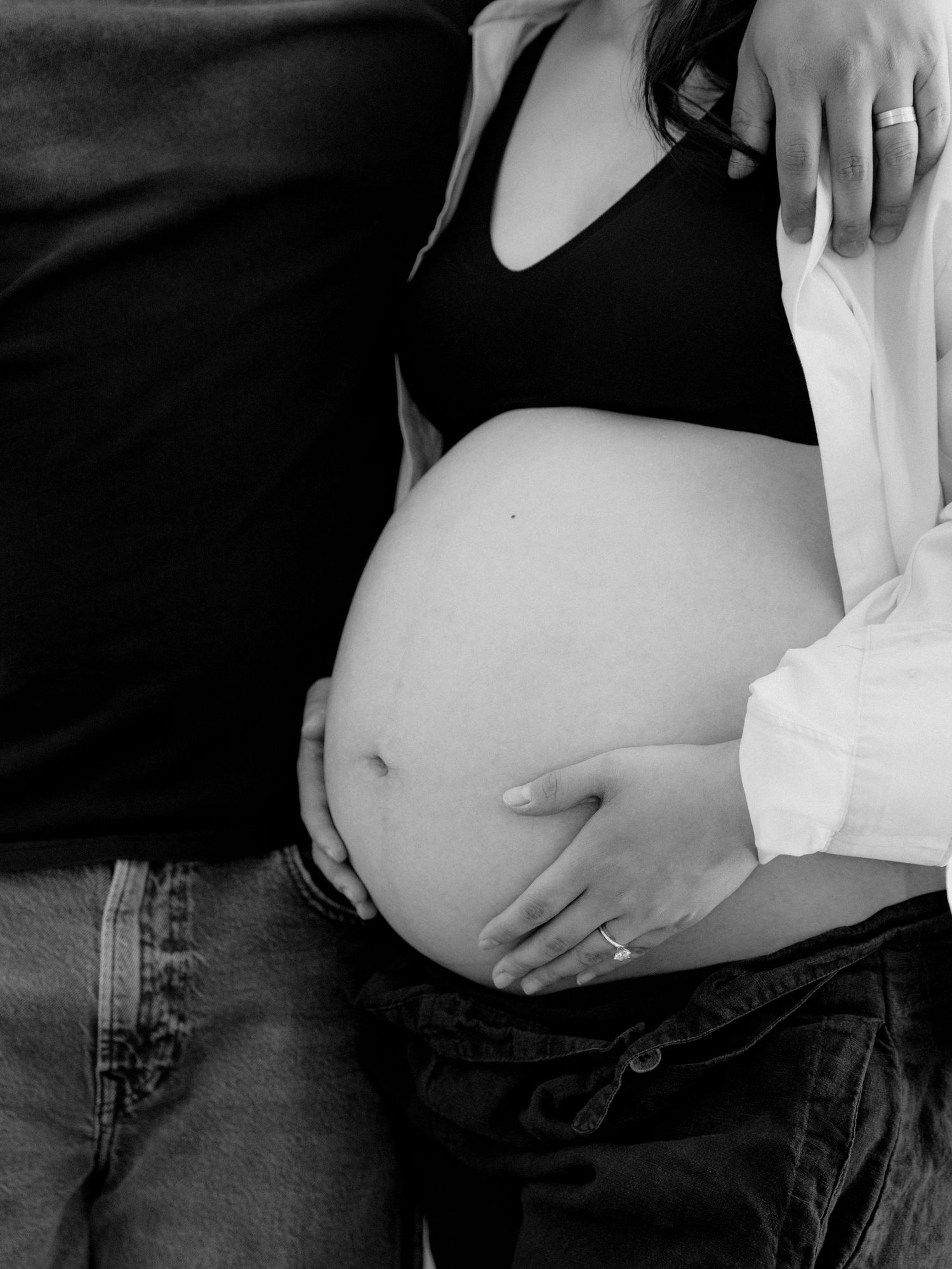Black and white maternity photo of pregnant woman's bare belly with partner's hand on shoulder and ring visible.