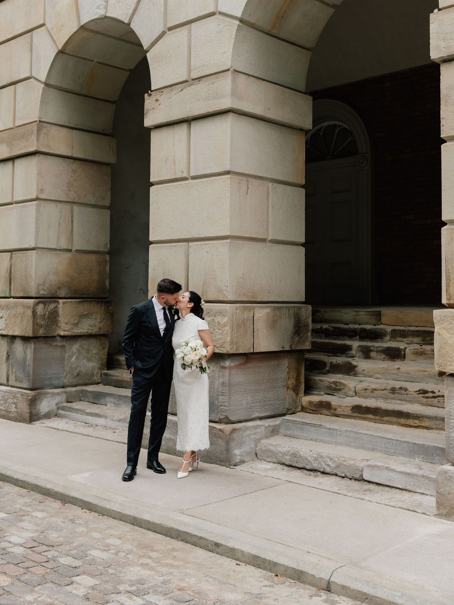 A couple shares a romantic kiss outside a grand stone building with arched columns on their wedding day.