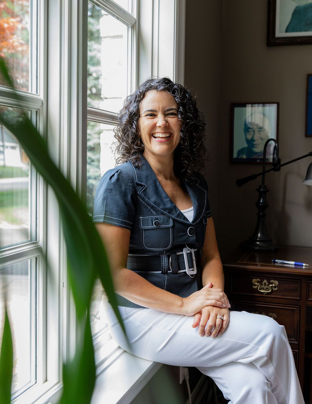 Smiling woman with curly hair sits by a bright window in a blue top and white pants in a cozy home office.
