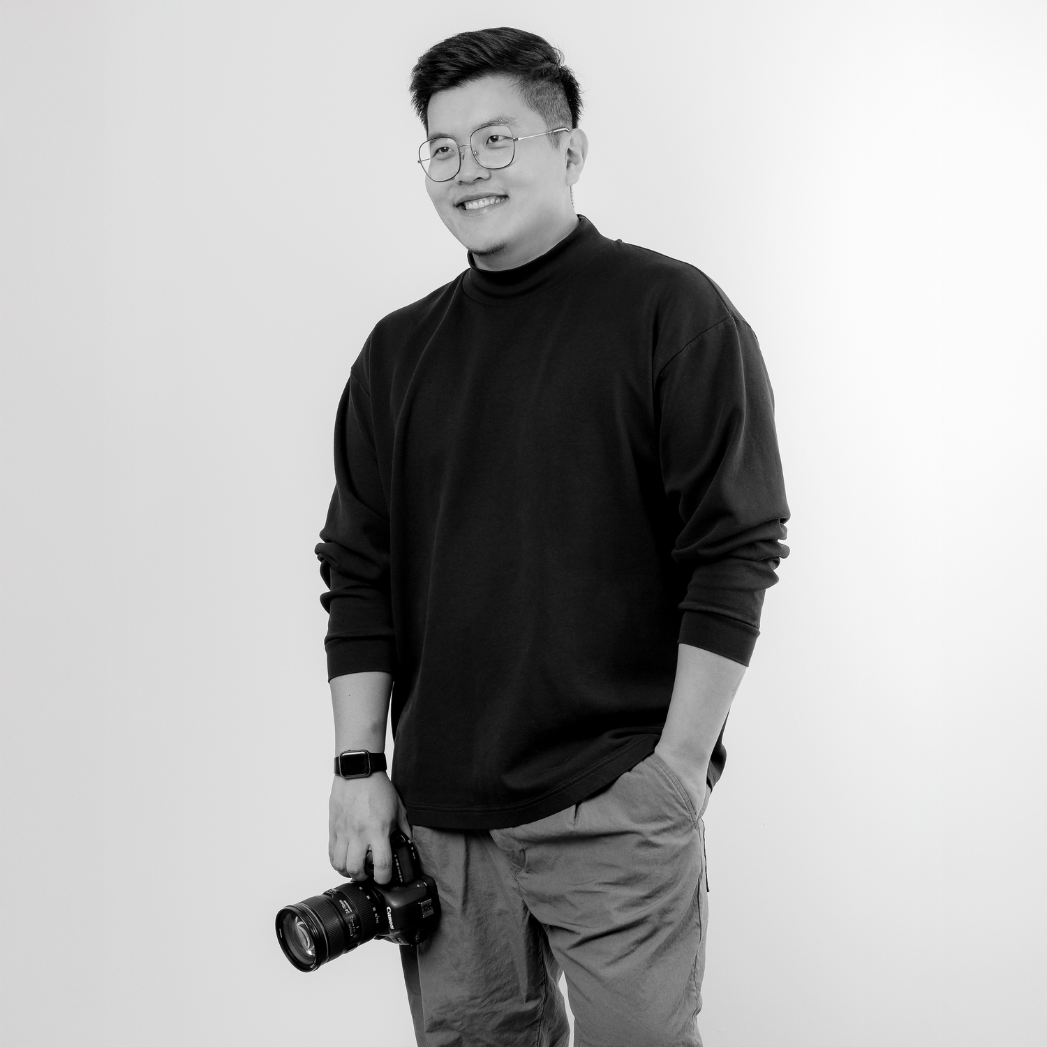 Black and white portrait of a smiling young man in a turtleneck holding a DSLR camera.