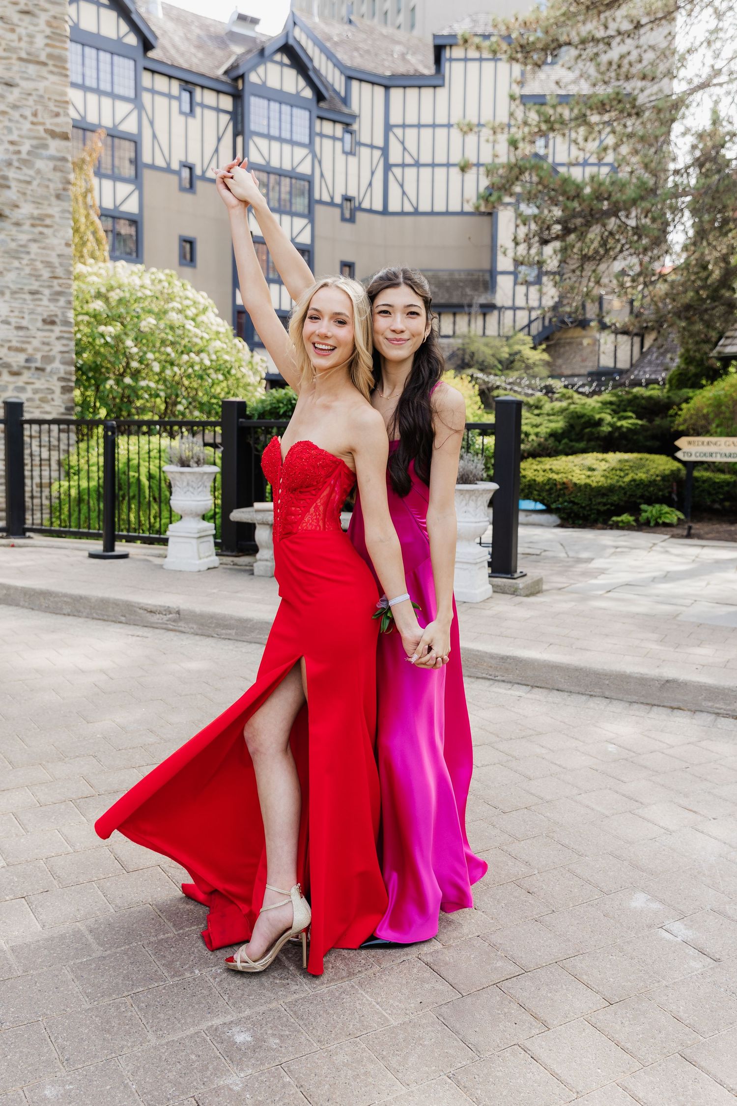 Two young women in elegant red and pink prom gowns posing outdoors near a stone building.