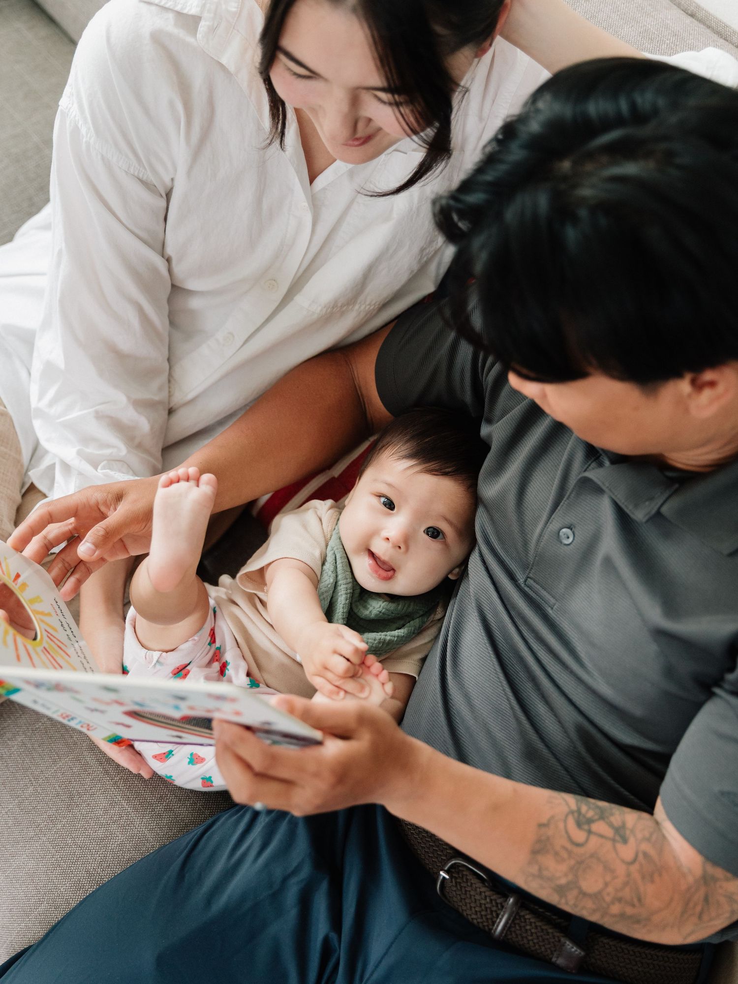 Parents reading a colorful picture book with their happy smiling baby on a cozy sofa at home.