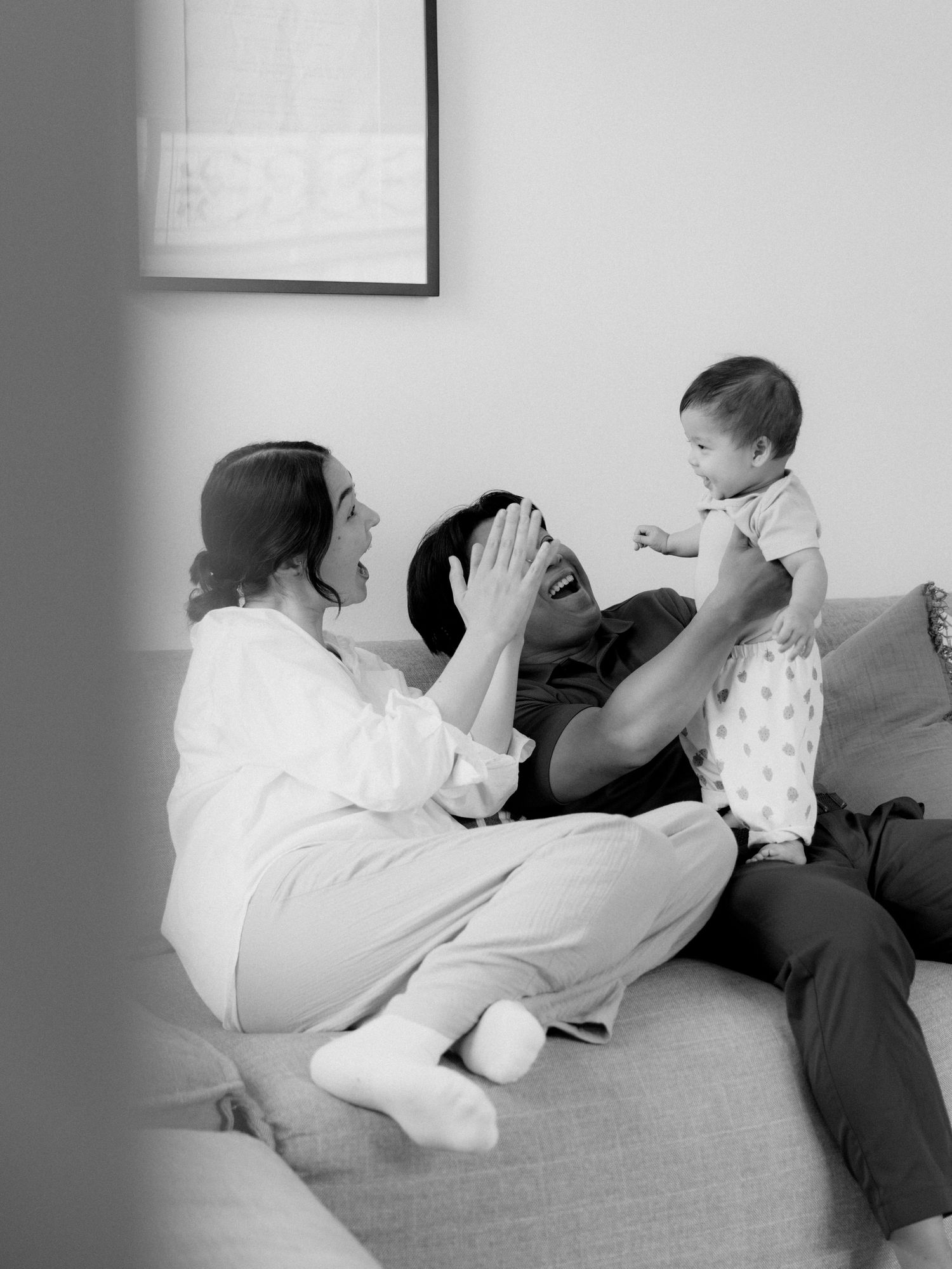 Black and white photo of two parents sitting on a couch, laughing while holding their baby up in the air.