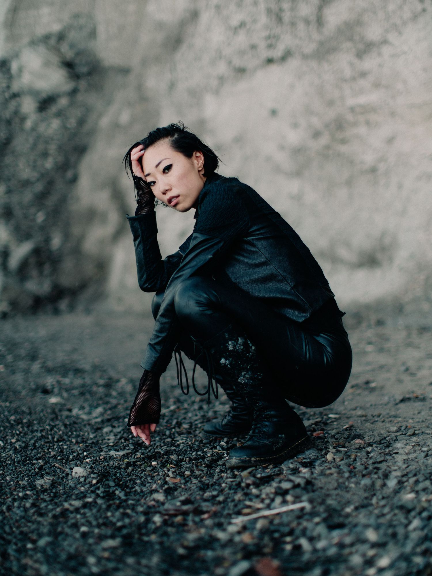Woman in black leather outfit crouching on rocky ground against a stone wall, looking pensively at camera.