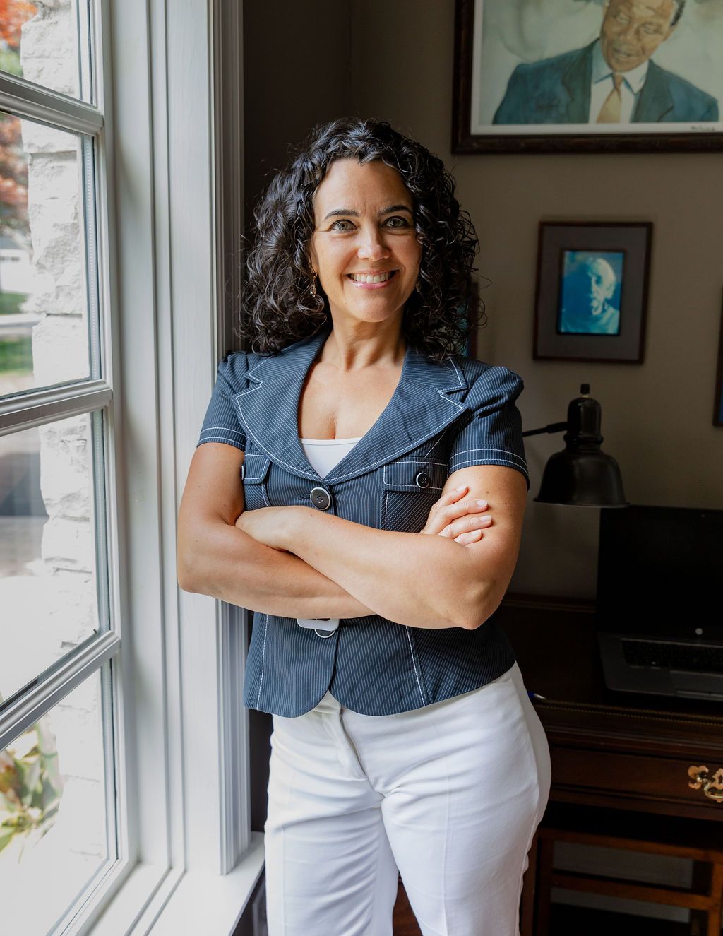 Professional woman in blue blazer and white pants smiling with arms crossed near a bright office window.